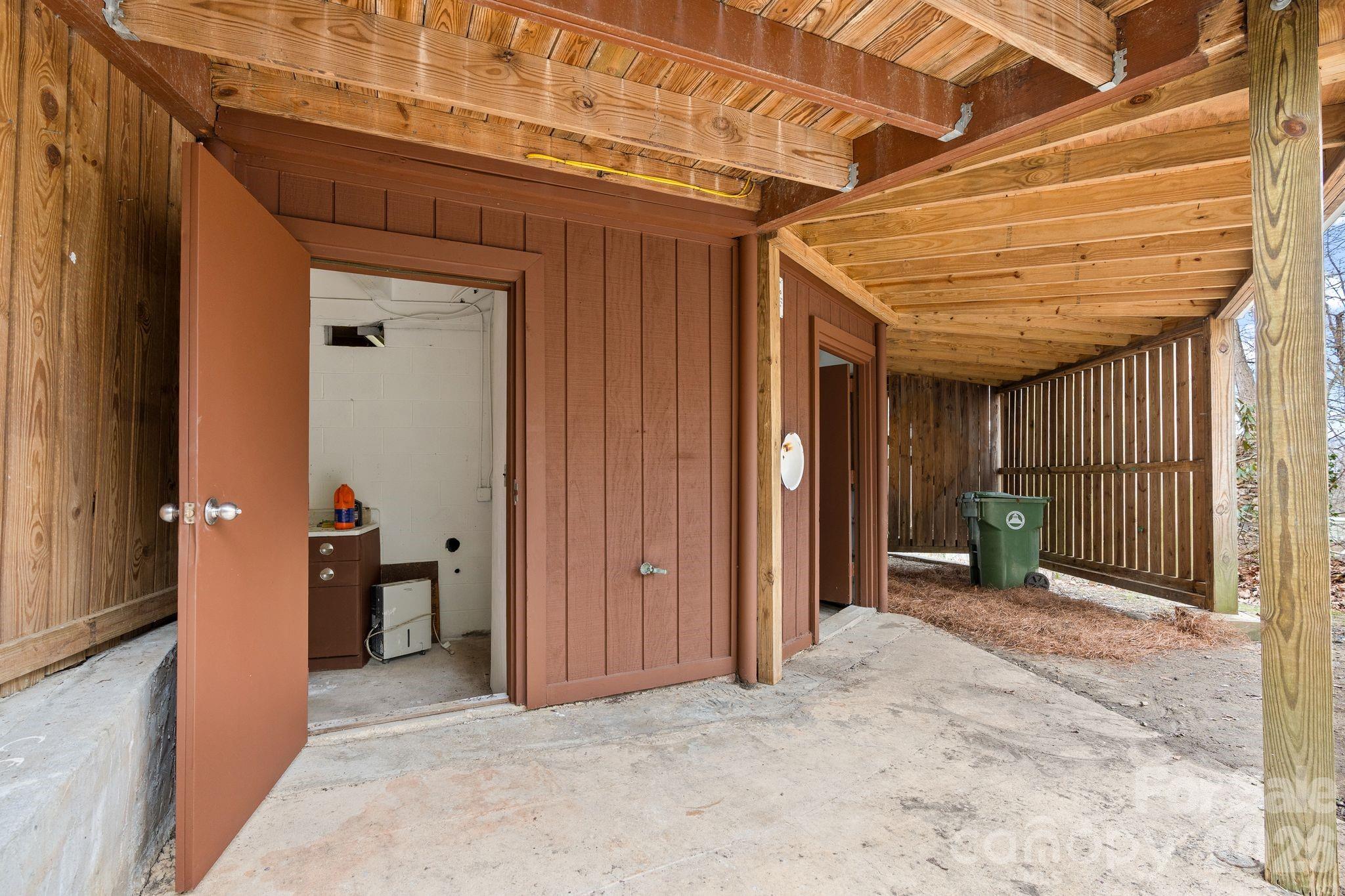 Covered outdoor utility area with exposed timber framing, concrete floor, and open storage-room doors.