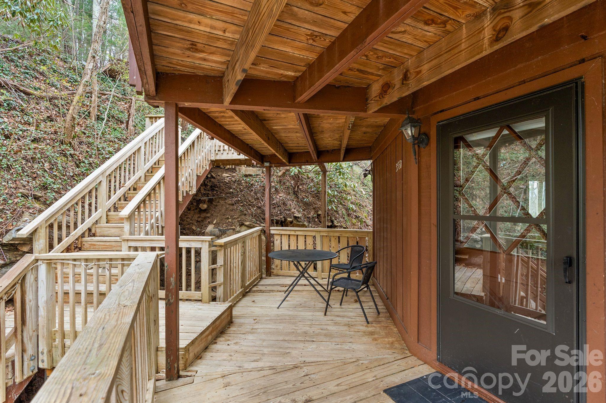 Covered wooden porch with black patio furniture and stairs climbing beside a wooded hillside.