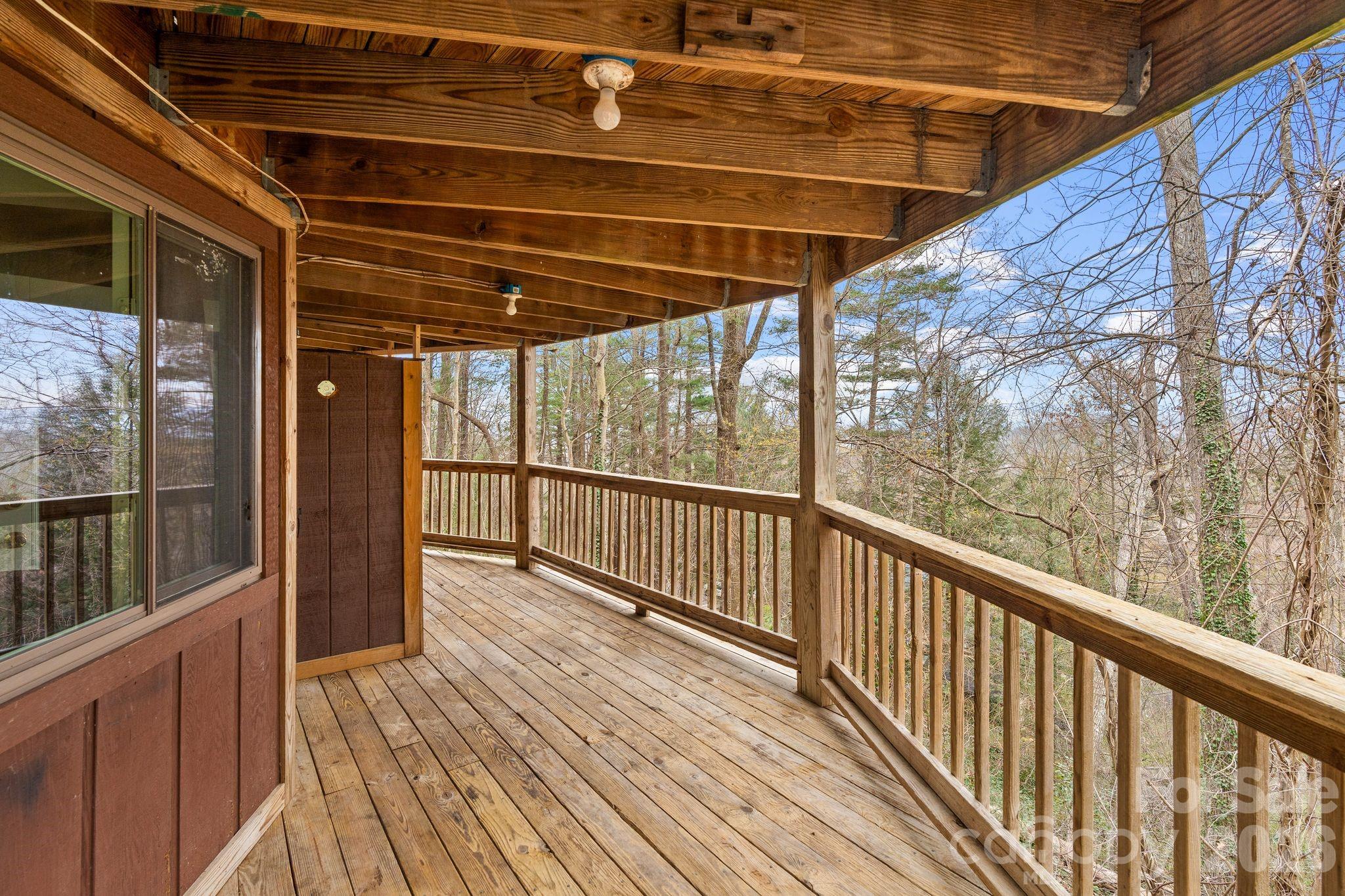 Covered wooden deck with exposed ceiling beams and a railing overlooking leafless trees.