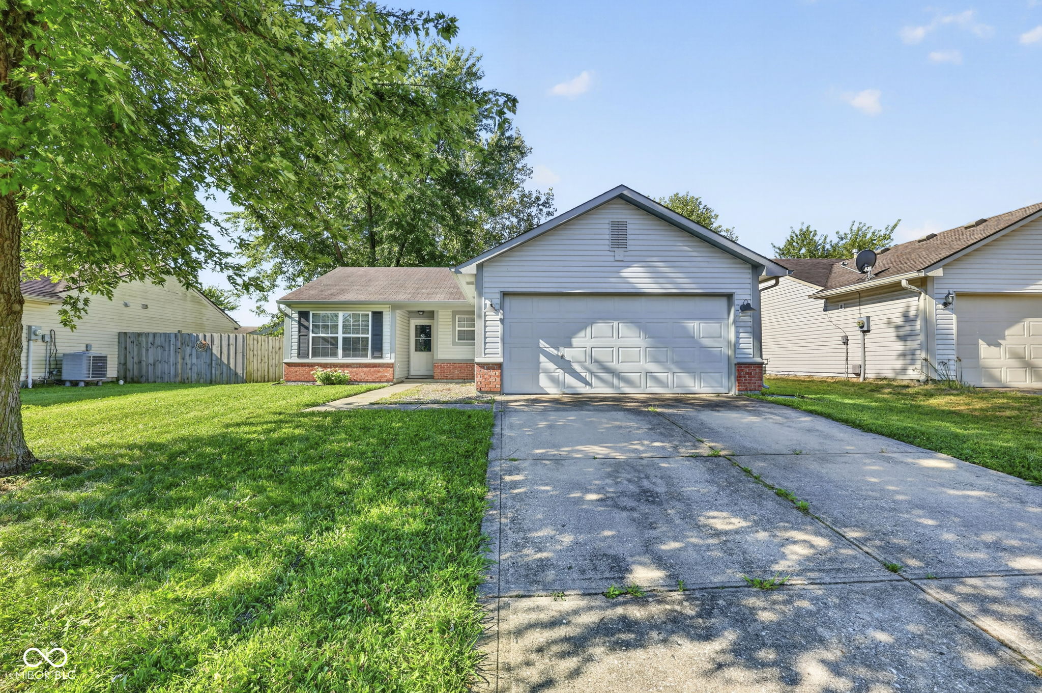 Single-story house with a two-car garage, concrete driveway, and large leafy tree shading the lawn.