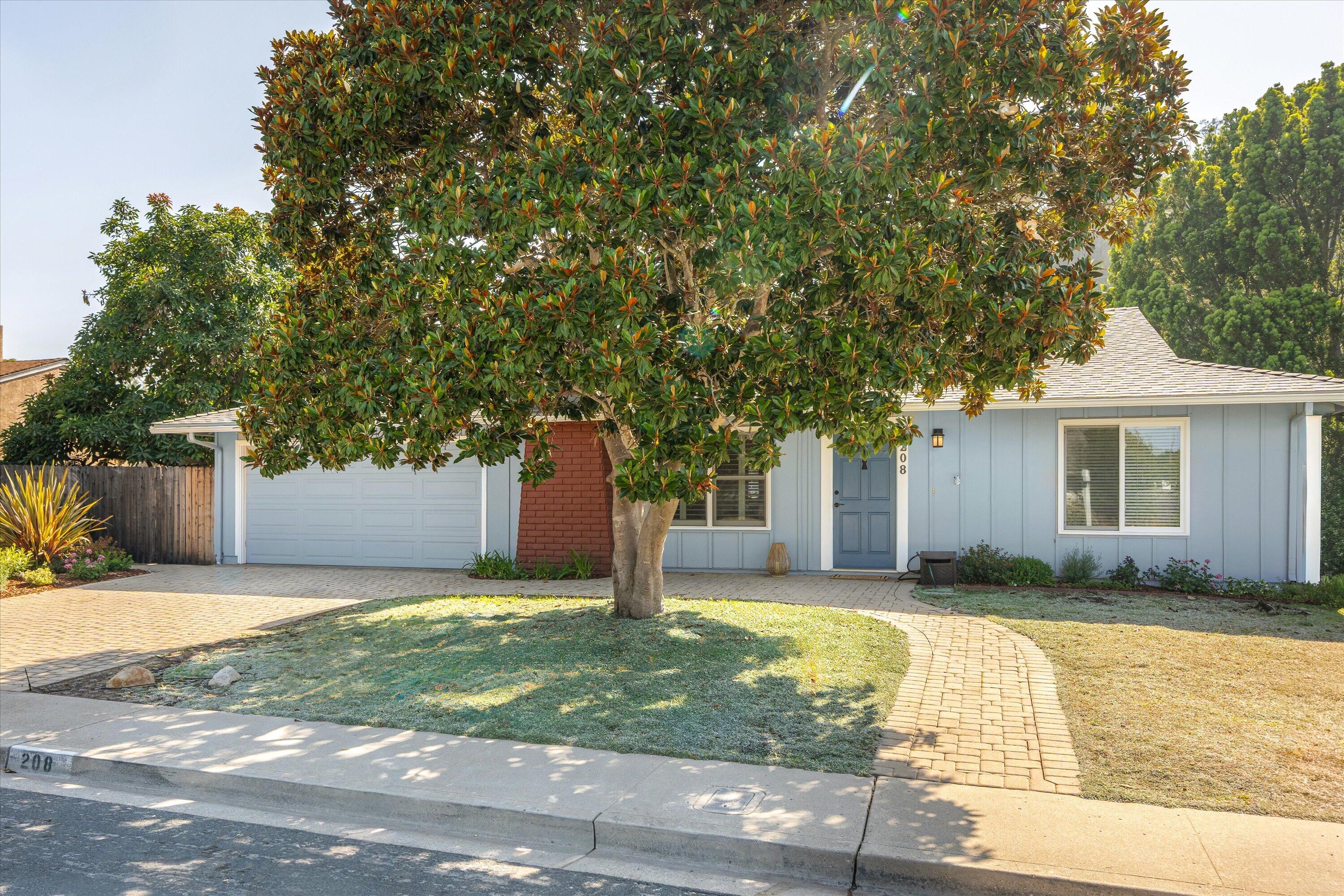 Light blue single-story house with a large shade tree, white garage door, and curved brick walkway.