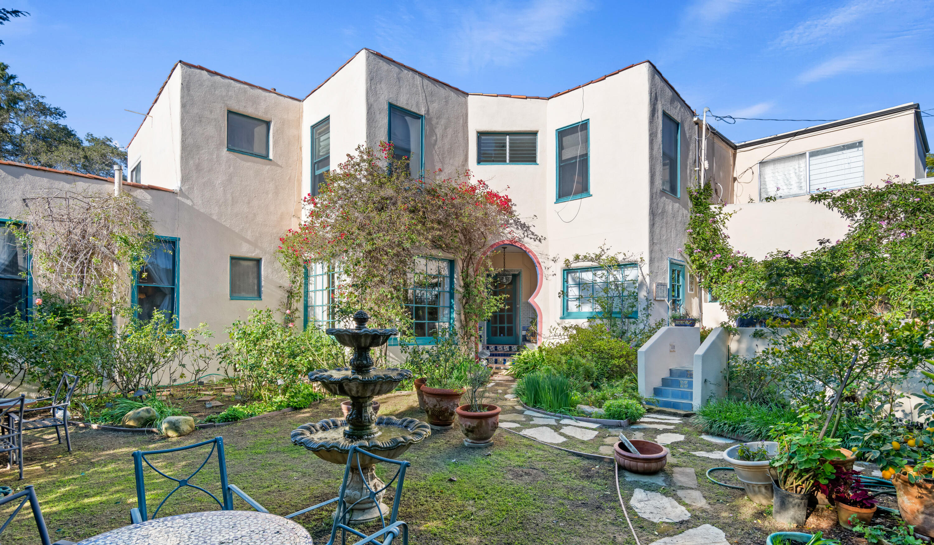 Two-story stucco home with an arched entry, teal-trimmed windows, and a lush garden with a tiered fountain.