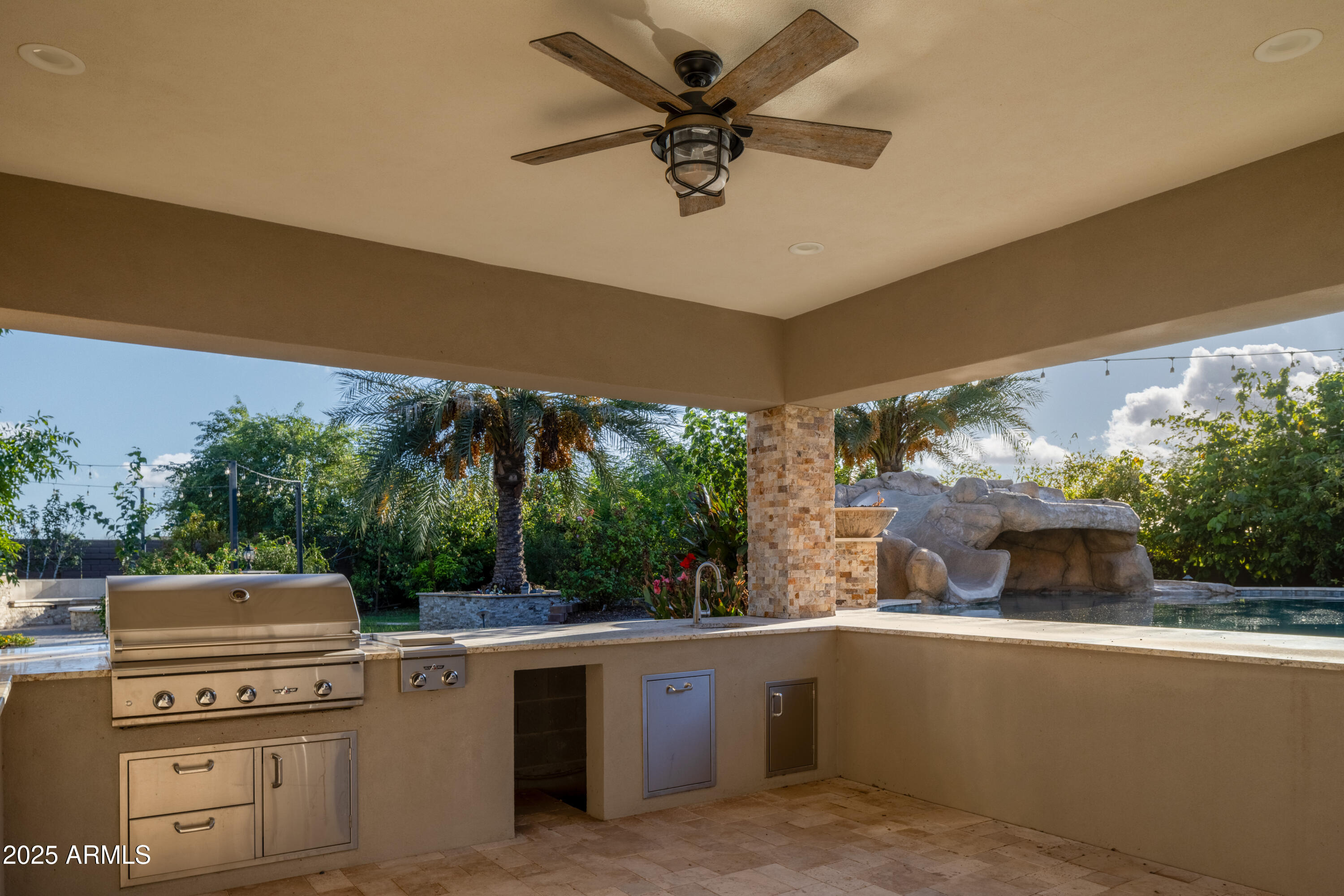 Covered outdoor kitchen with a built-in grill overlooking a pool and rock waterfall.