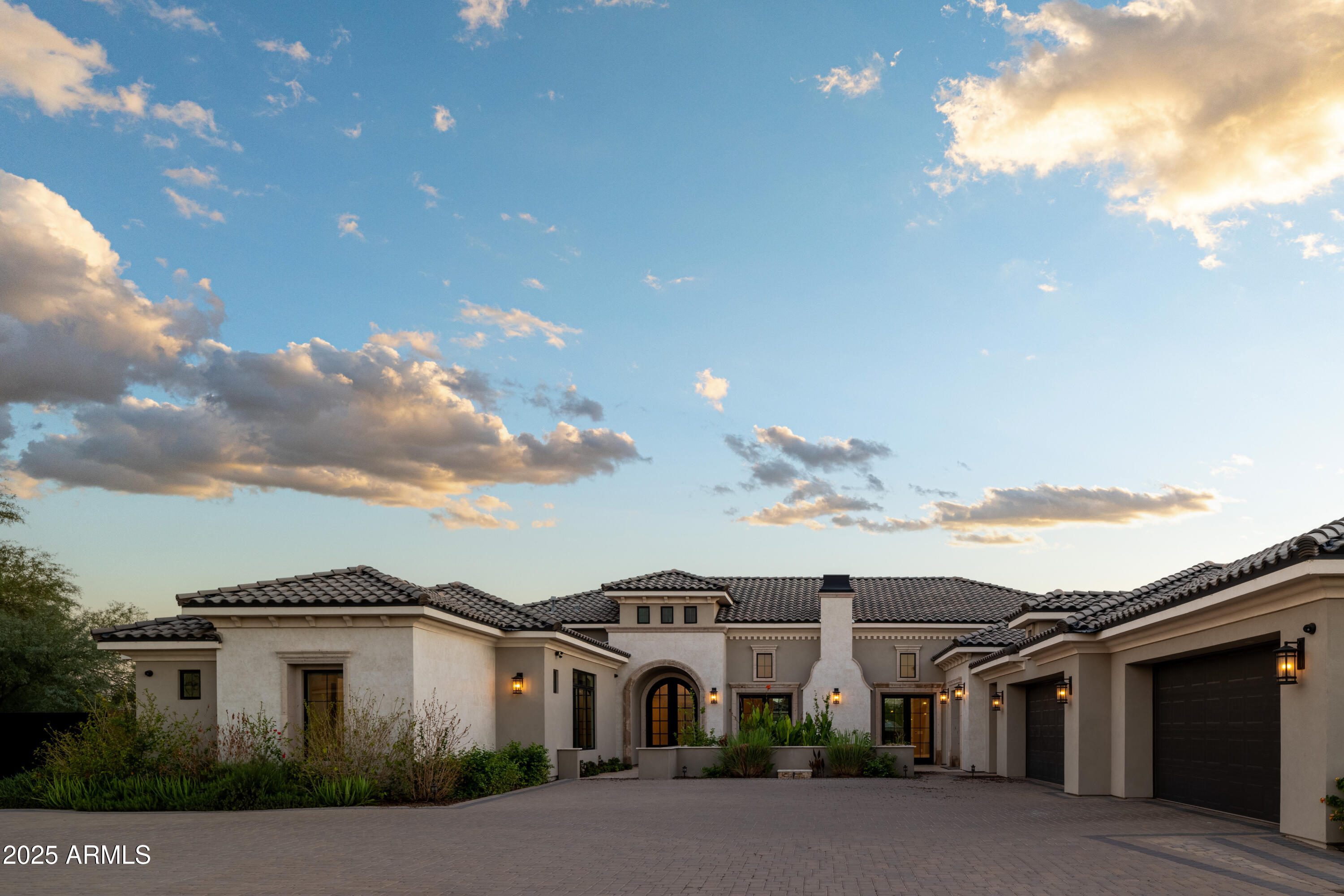 Large stucco home with tiled roof, arched entry, landscaped courtyard, and attached garages at sunset.