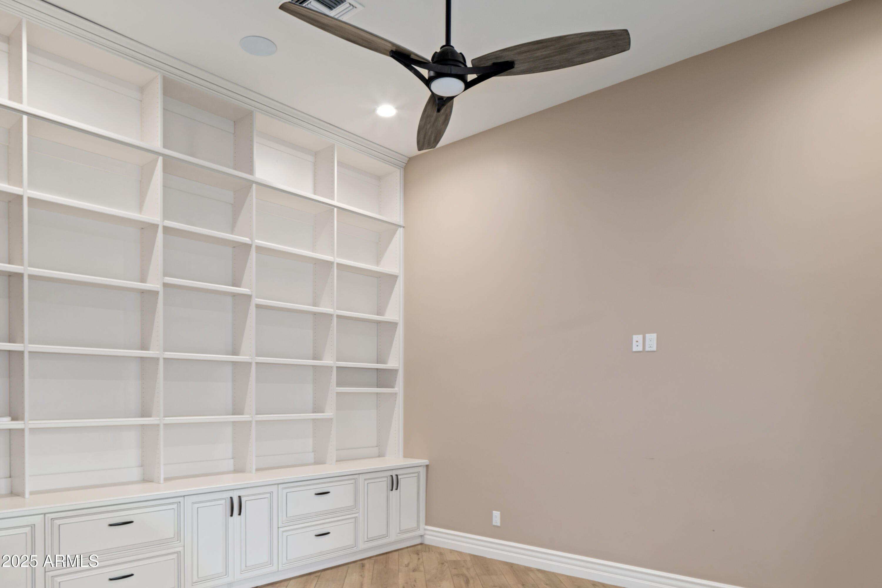White built-in bookshelves and cabinets line a room with tan walls and light wood flooring.