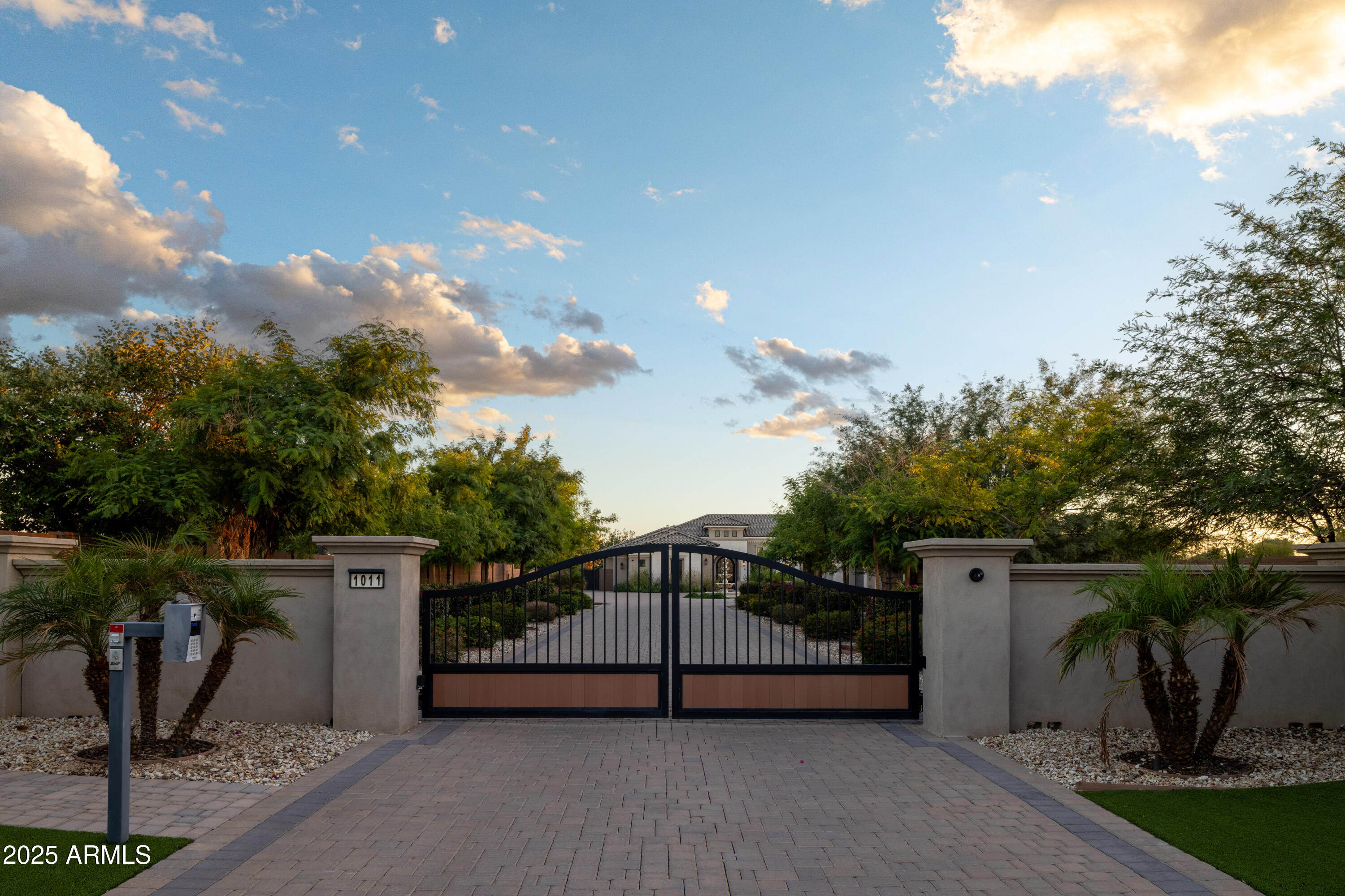 Gated home entrance with a black metal driveway gate, landscaped stucco walls, and a paved drive.