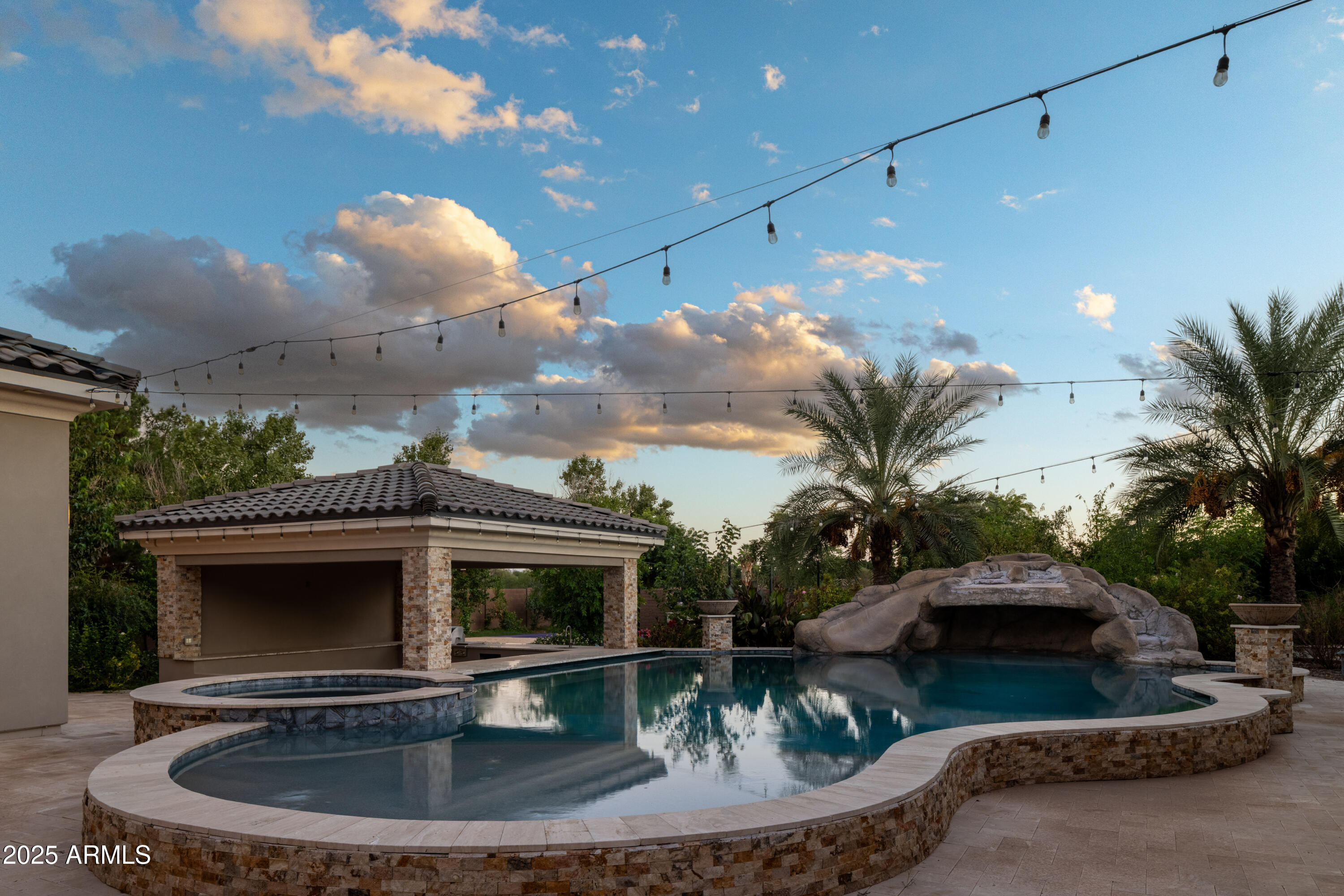 Backyard pool and raised spa with a rock waterfall, pavilion, palm trees, and overhead string lights.