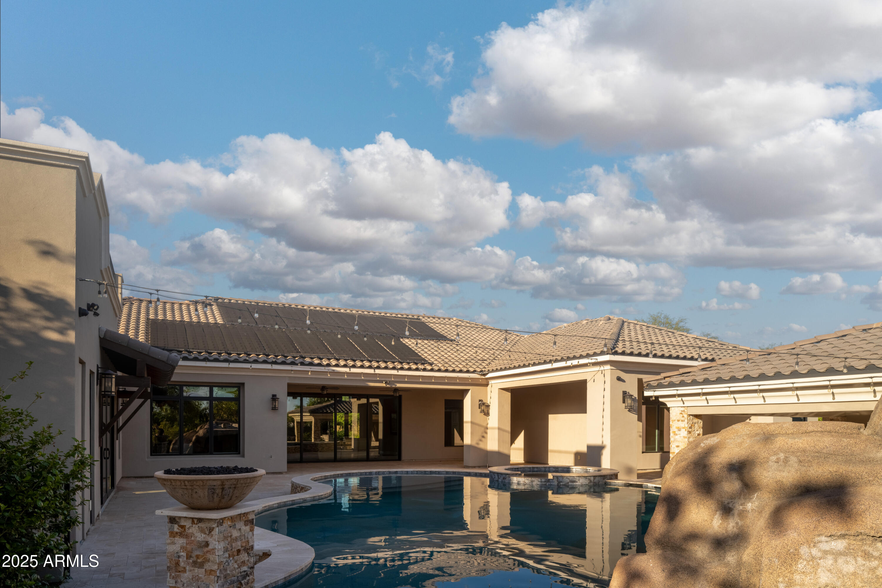 Home’s rear courtyard with a curved pool, raised spa, covered patio, and large decorative boulder.