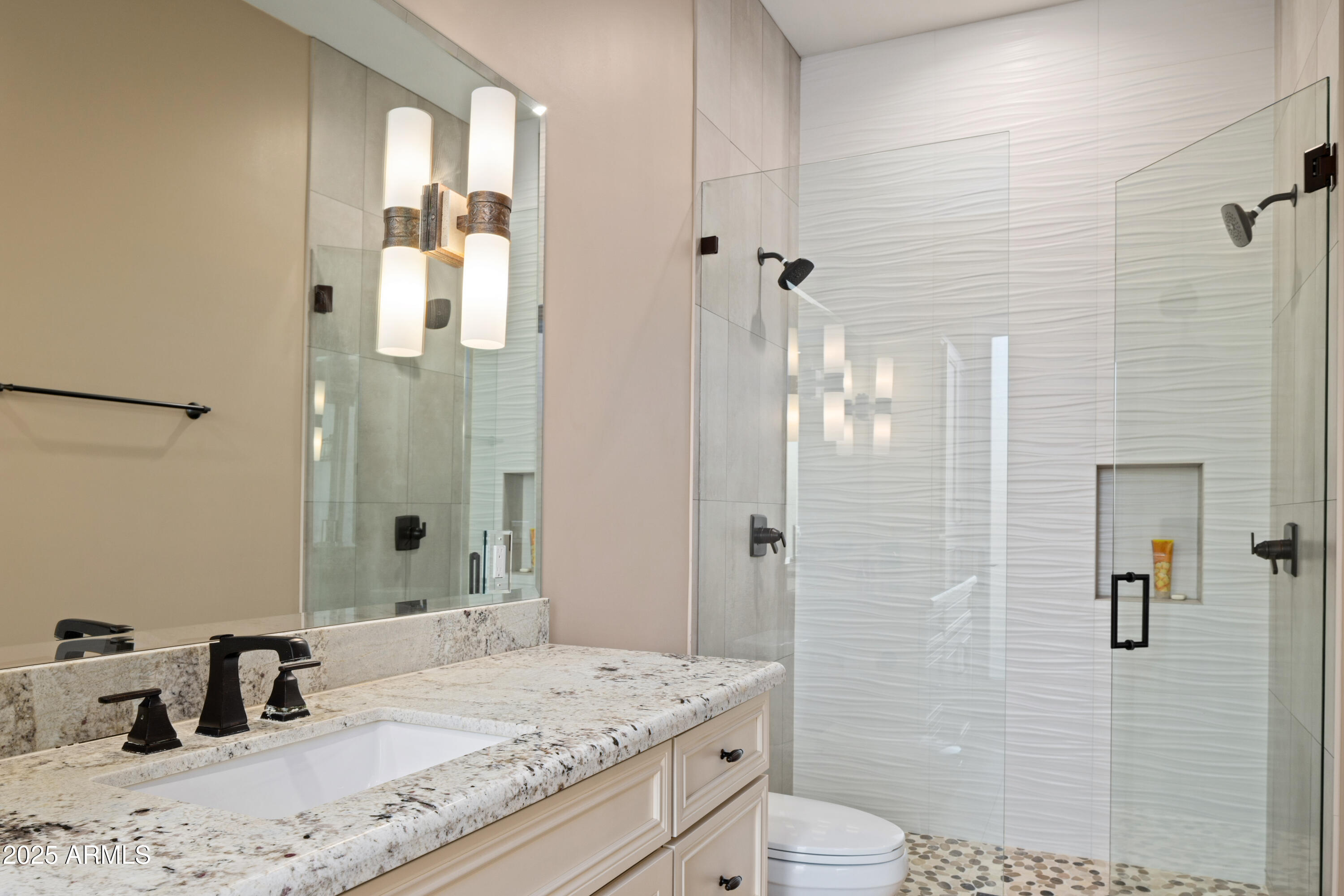Bathroom with granite vanity and glass shower featuring wavy white tile and pebble flooring.