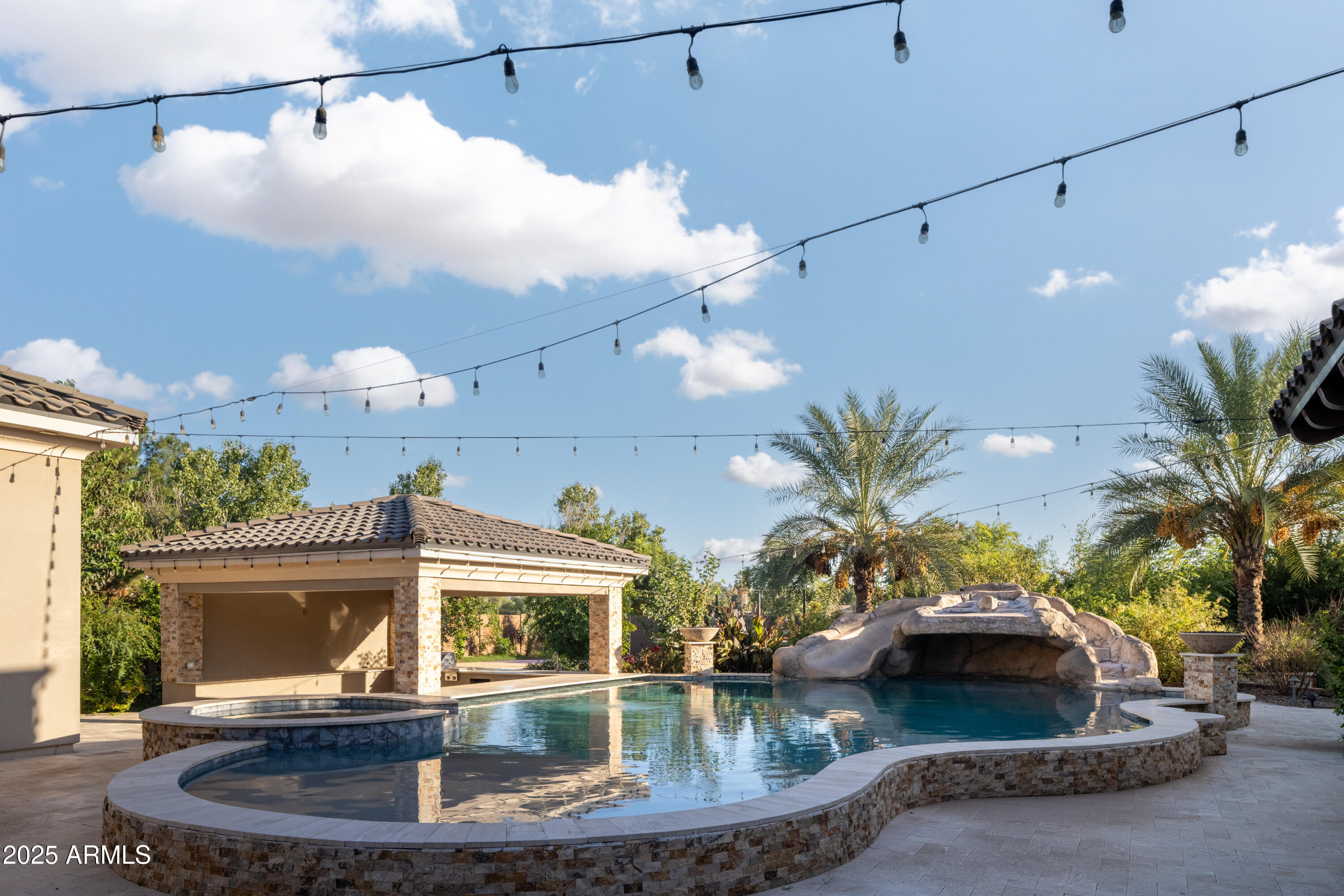 Backyard pool with a rock waterfall, attached spa, tiled-roof pavilion, and overhead string lights.