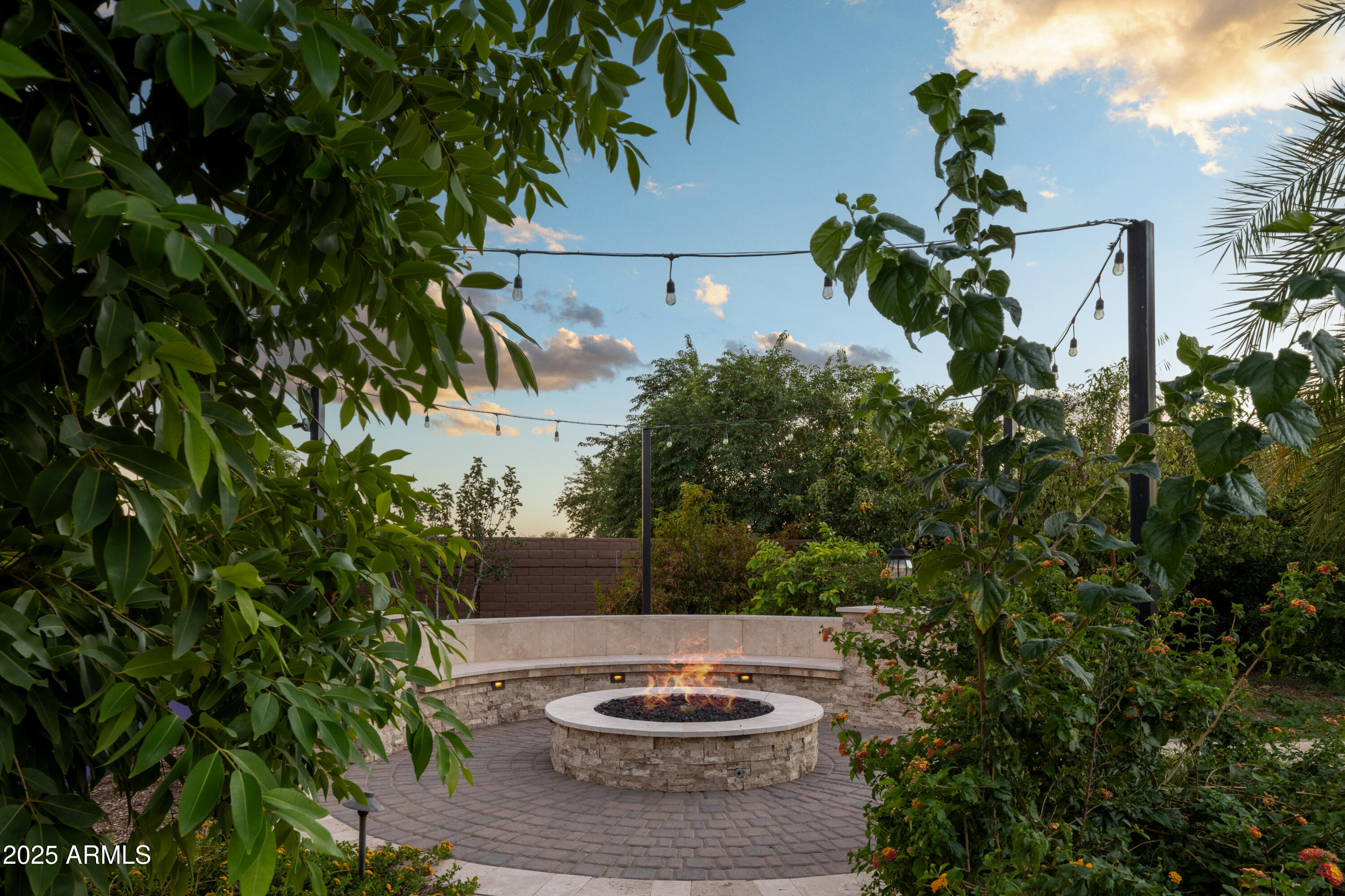 Backyard patio with a circular stone fire pit, curved seating wall, and overhead string lights.