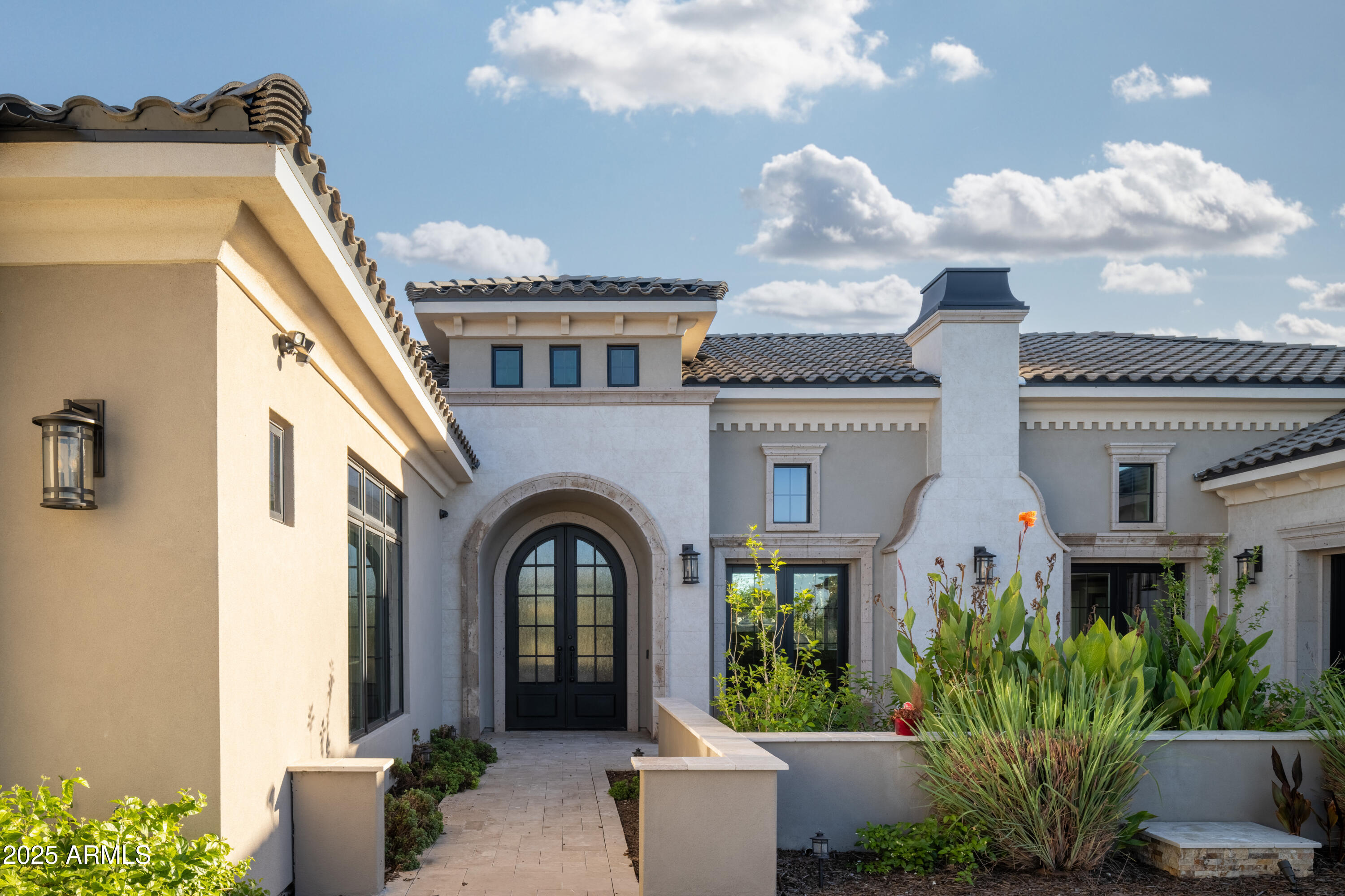 Stucco home entrance with an arched black double door and landscaped courtyard.