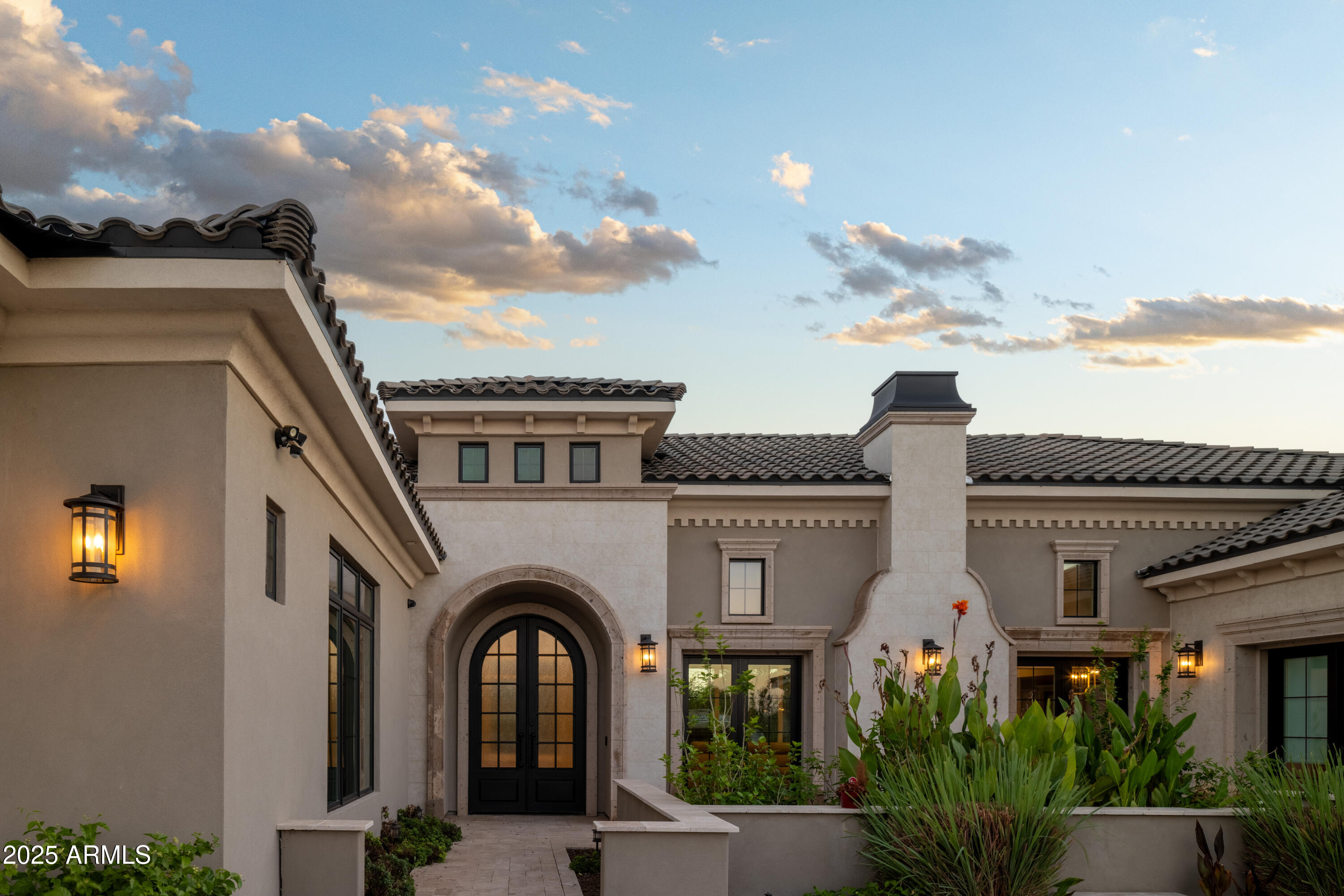 Home exterior with an arched black entry, tiled roof, and landscaped courtyard at dusk.