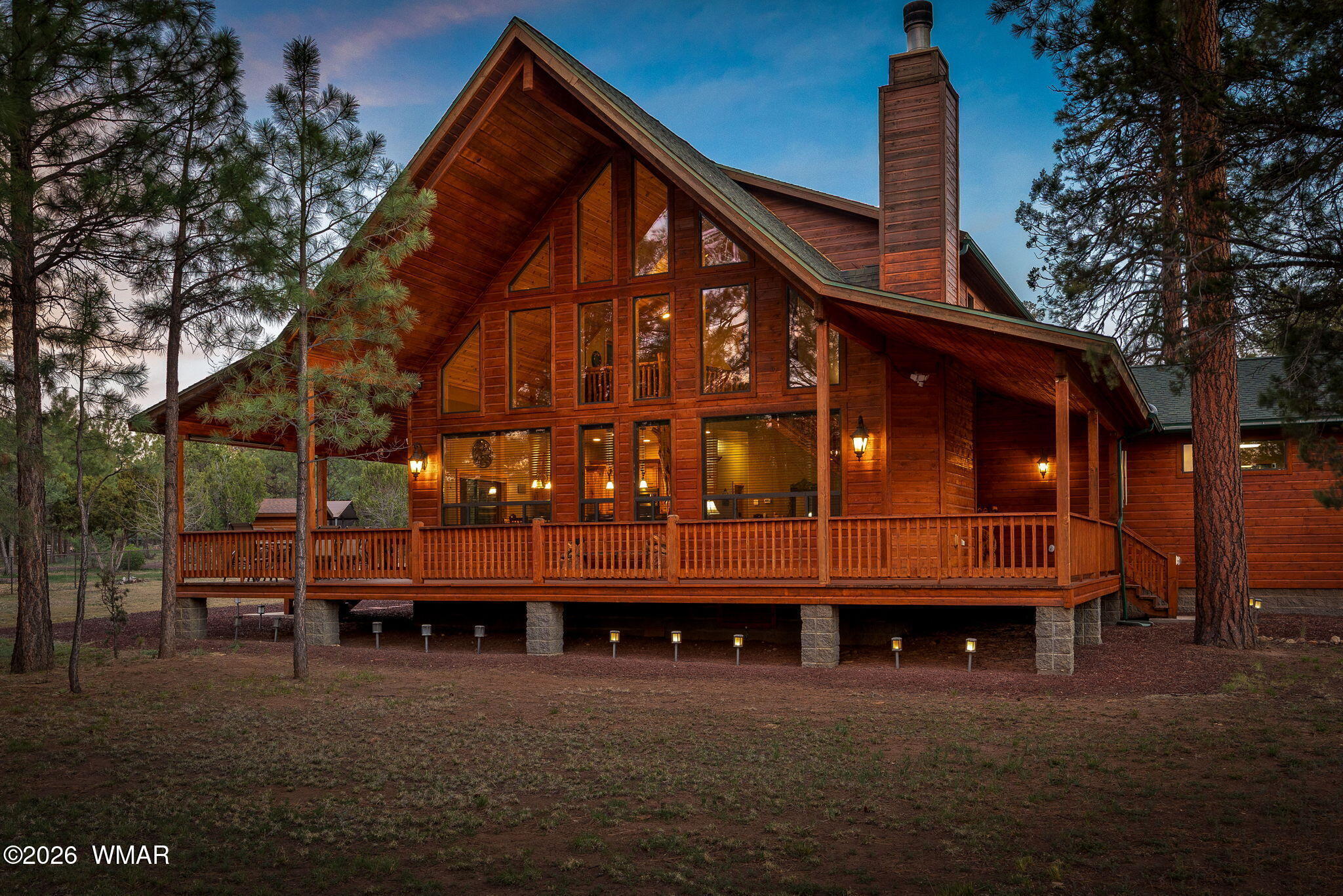 Wooden lodge with tall windows and a raised wraparound deck among pine trees.