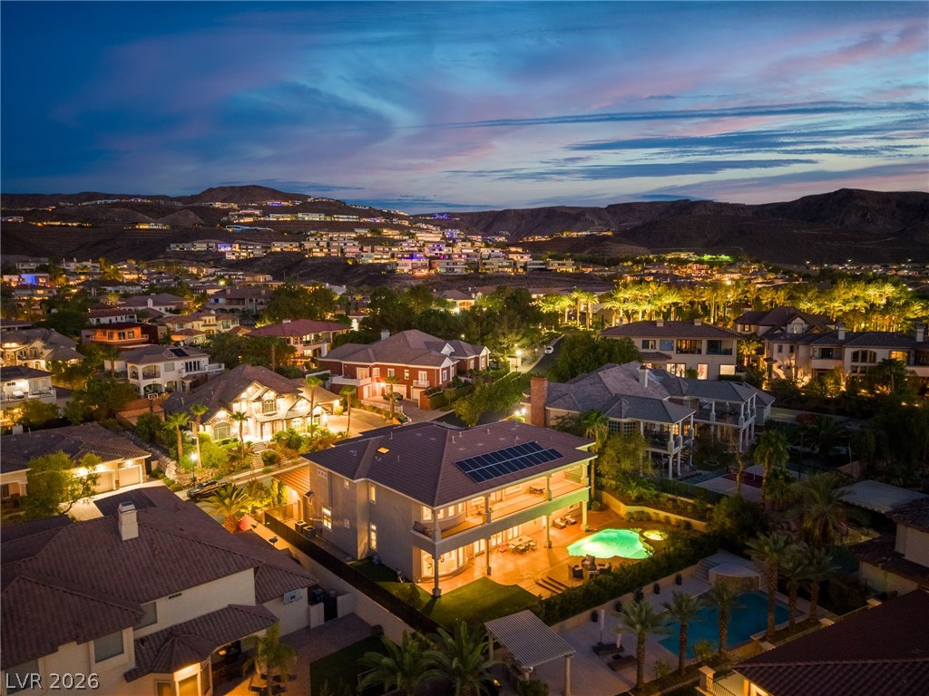 Aerial view of a two-story home with rooftop solar panels and an illuminated pool, surrounded by hillside homes at dusk.