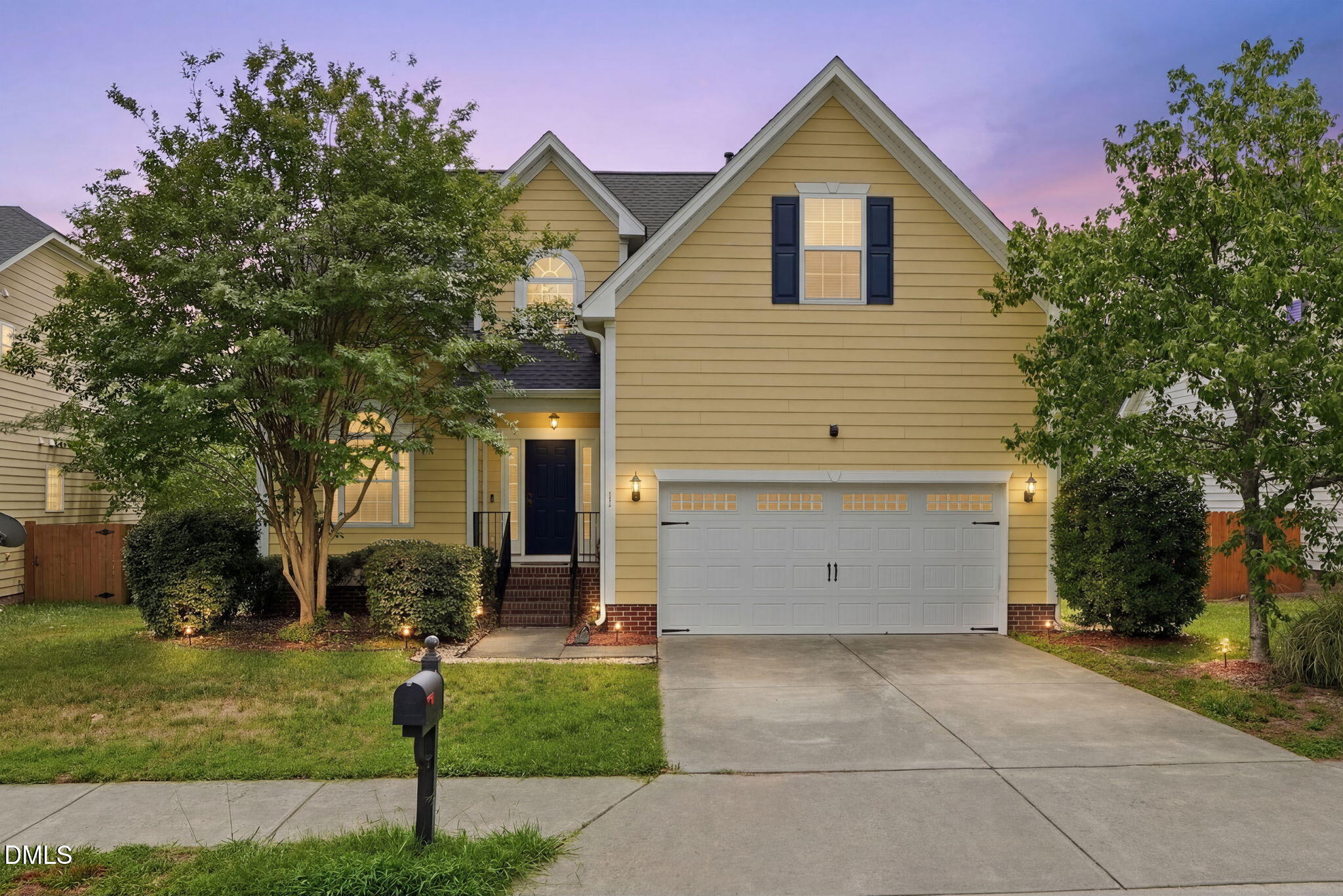 Yellow two-story home with dark shutters, illuminated windows, double garage, and landscaped front yard.