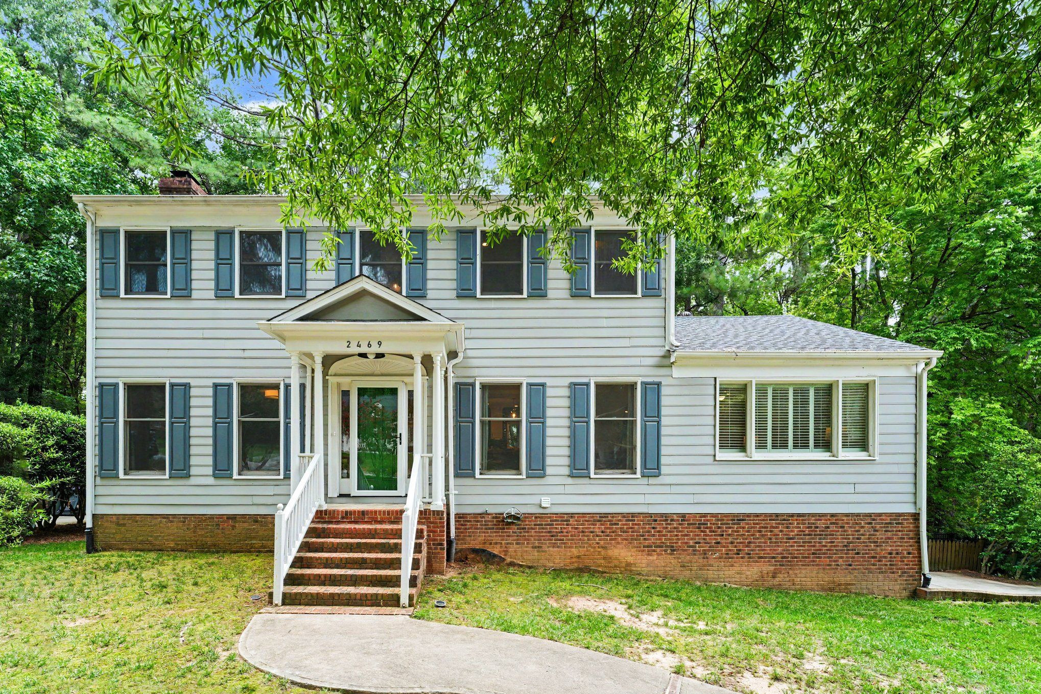 Two-story house with blue shutters, a columned porch, and brick front steps beneath leafy trees.