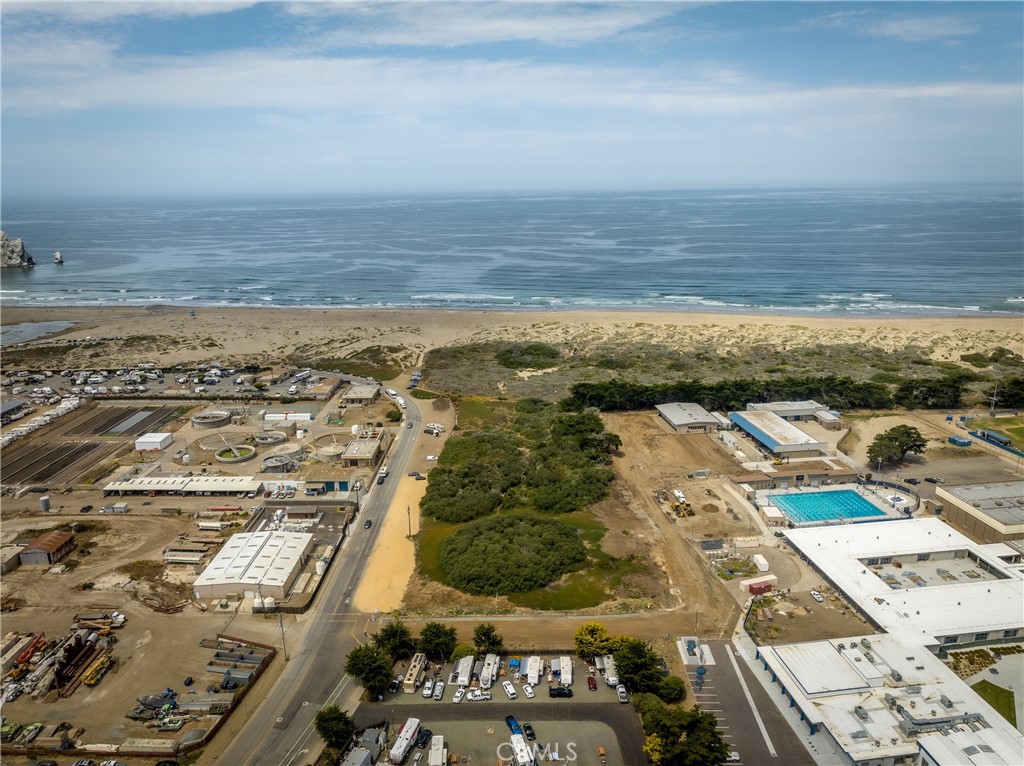 Vacant coastal parcel with low vegetation, a road, beach, and ocean beyond.