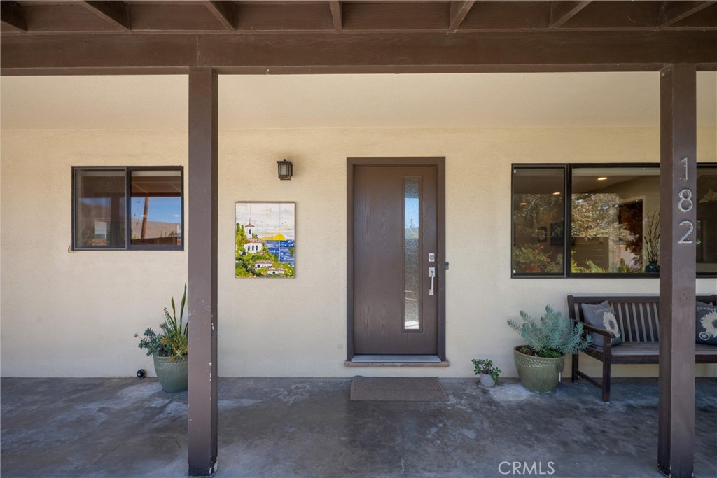 Covered front porch with a dark entry door, wall art, potted plants, and a wooden bench.