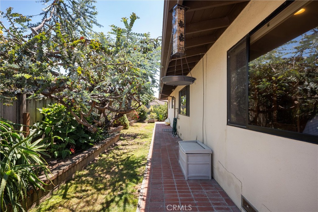 House side yard with a red tile walkway beside mature trees and planted garden beds.