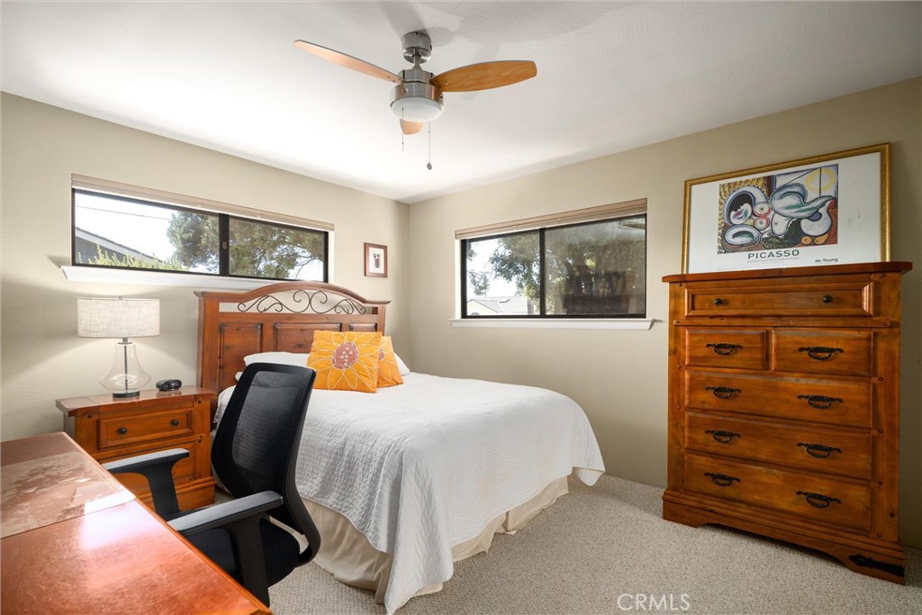 Furnished bedroom with a white-covered bed, wooden furniture, two windows, and a ceiling fan.
