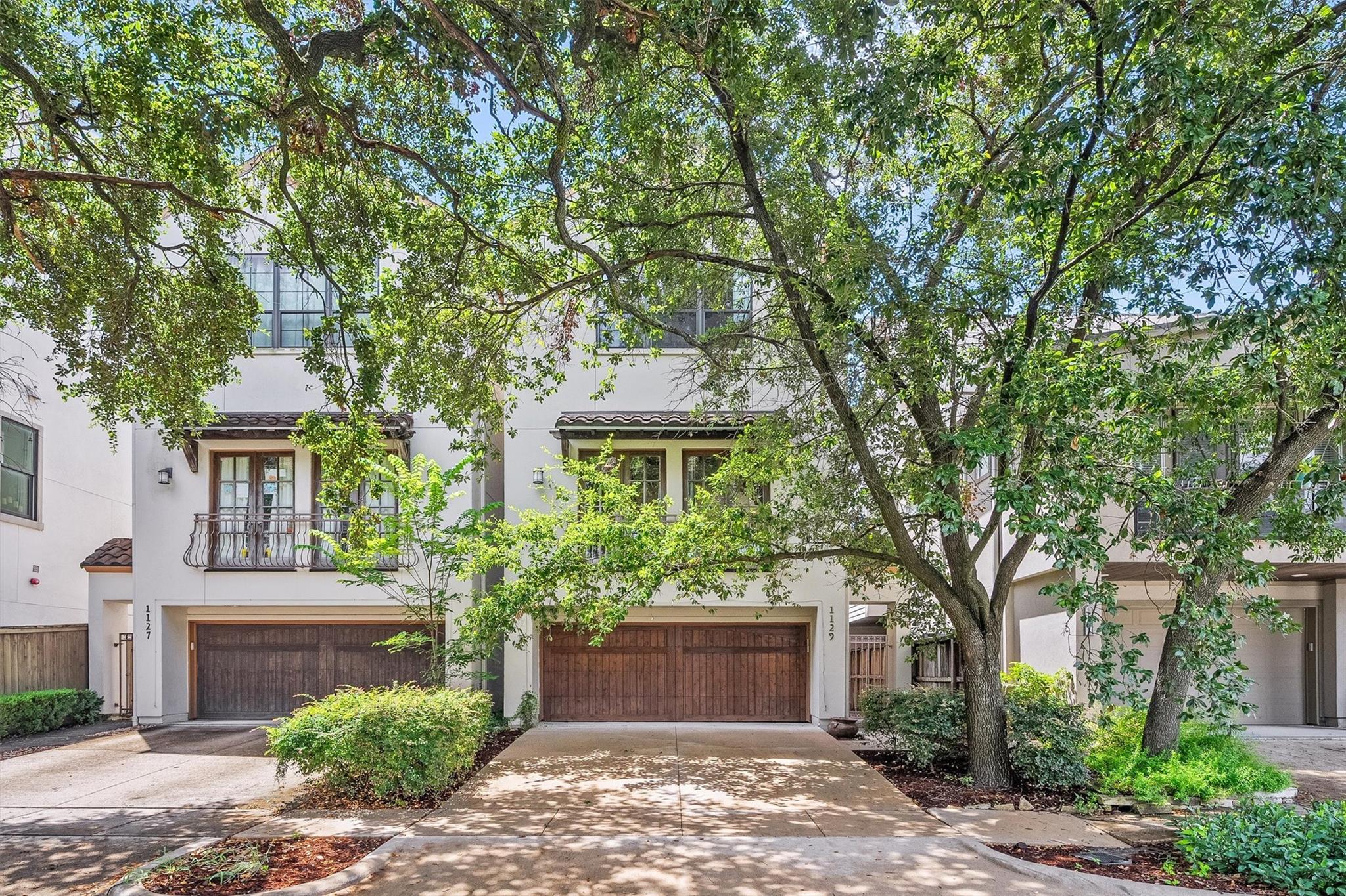 Townhouse with a wood-paneled garage door and balcony beneath mature shade trees.