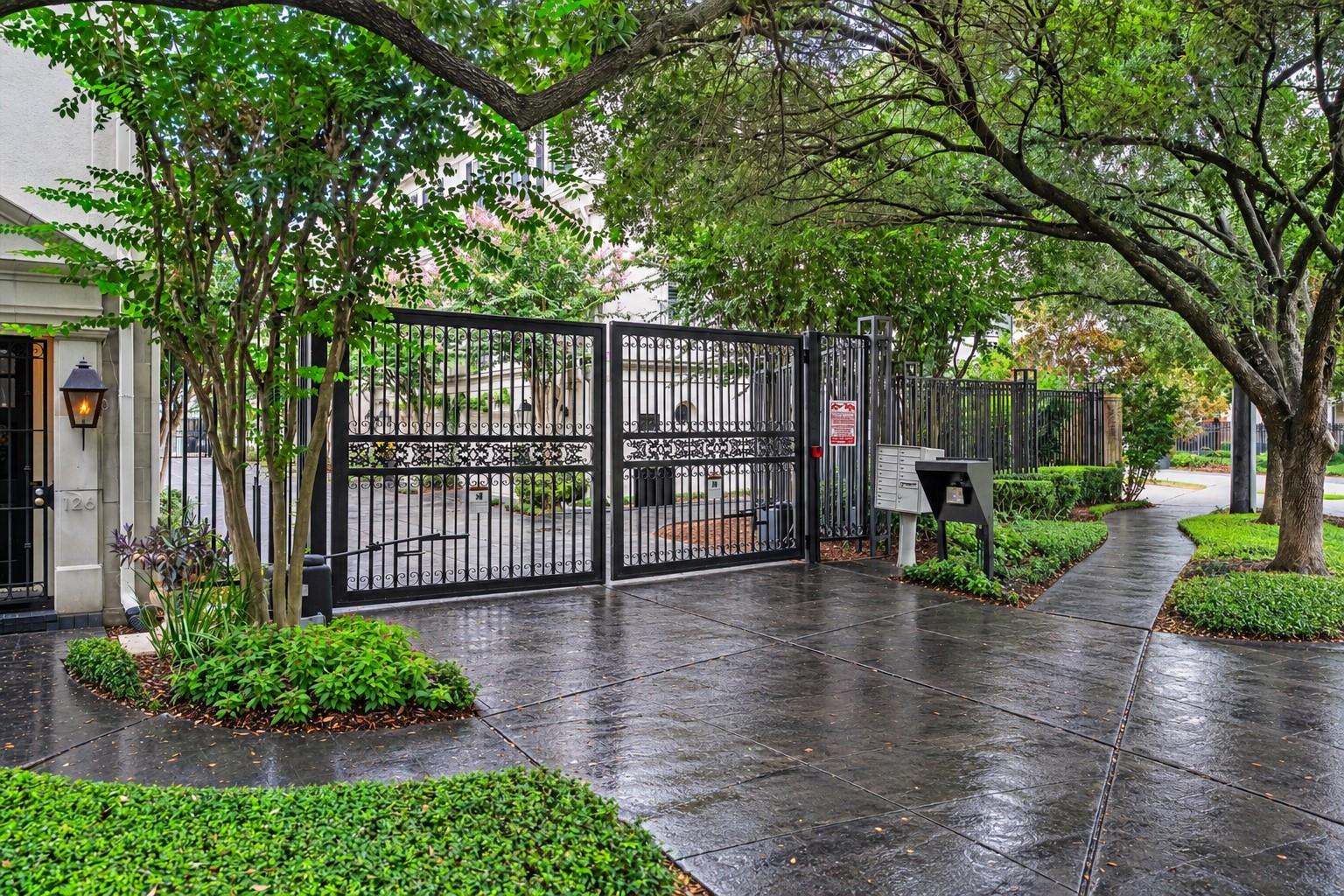 Residential entrance with ornate black gates and a tree-lined walkway wet from rain.