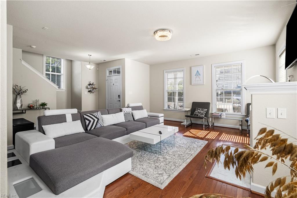Living room with a gray sectional, patterned rug, hardwood floors, and tall windows with blinds.