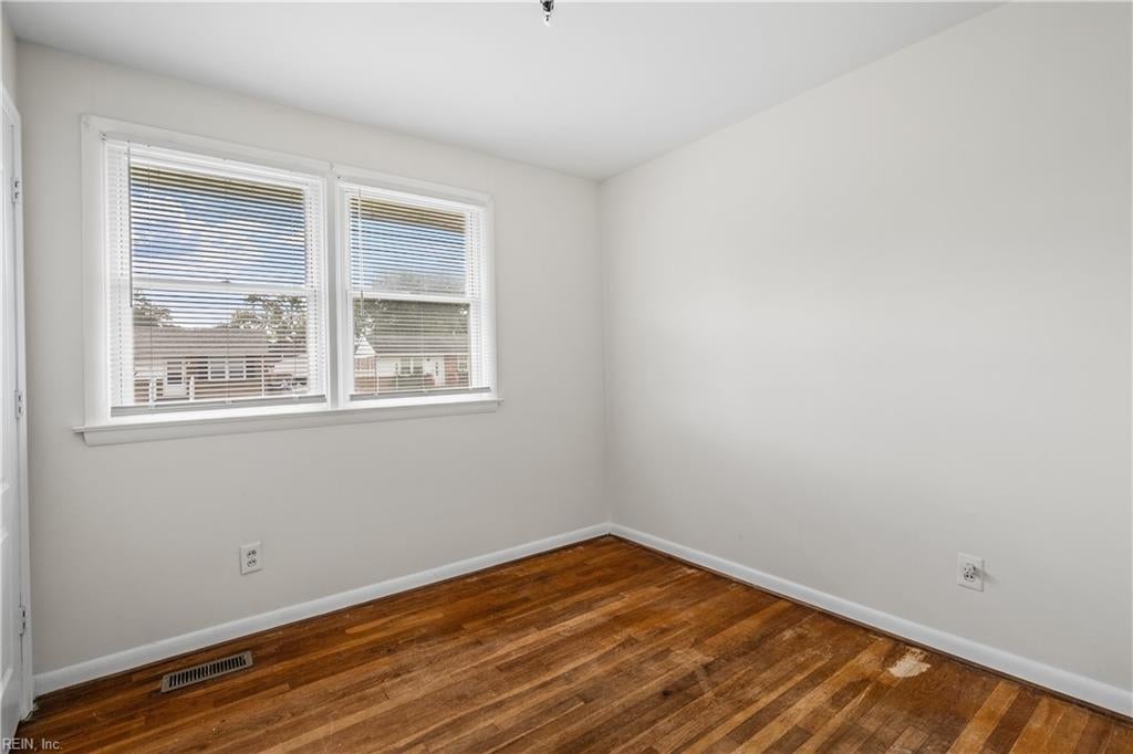 Empty room with hardwood floors, two windows with blinds, and a floor vent.