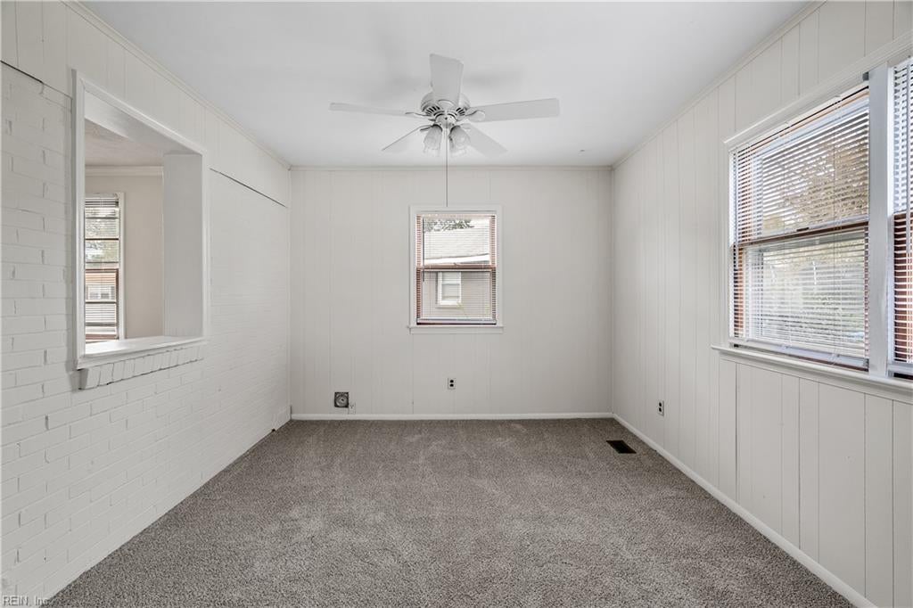 Empty carpeted room with white paneled walls, a brick pass-through, multiple windows, and a ceiling fan.