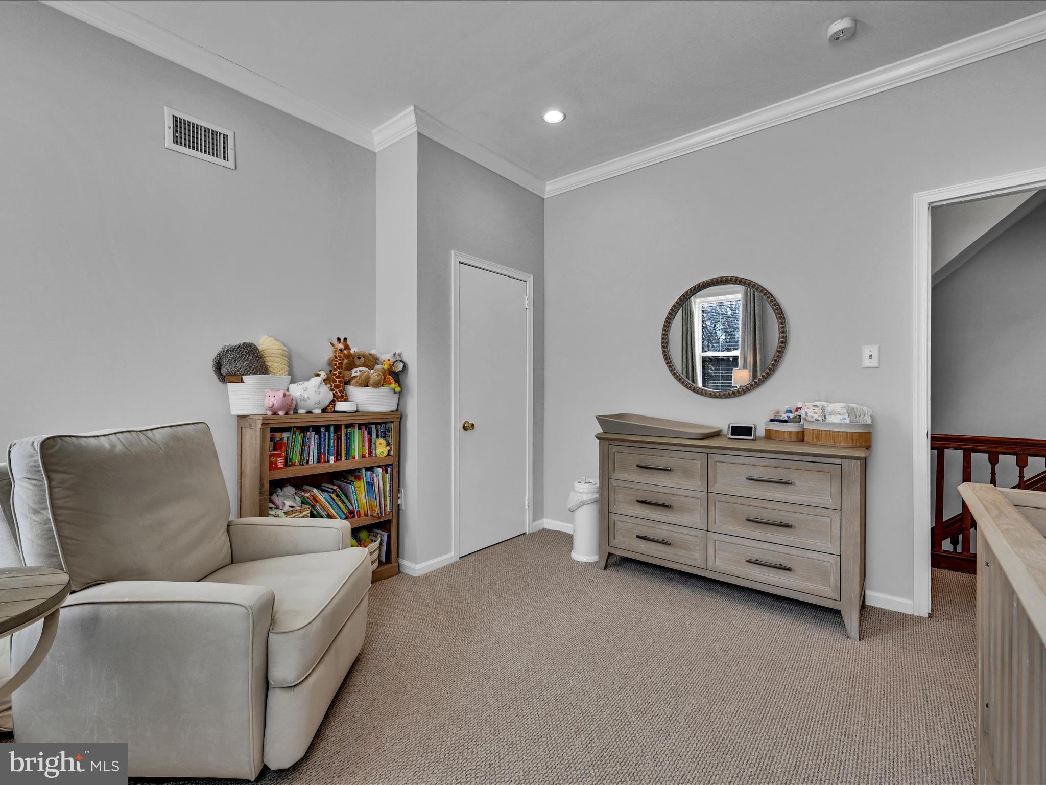 Furnished nursery with a glider, children’s bookcase, and wooden dresser beneath a round mirror.