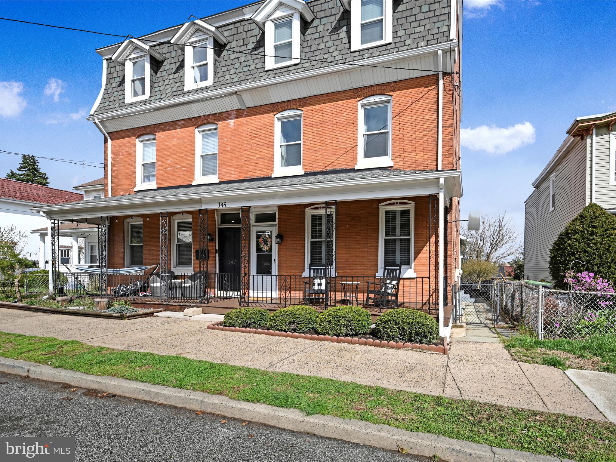 Red-brick home with gray mansard roof and a covered porch lined with iron railings and seating.