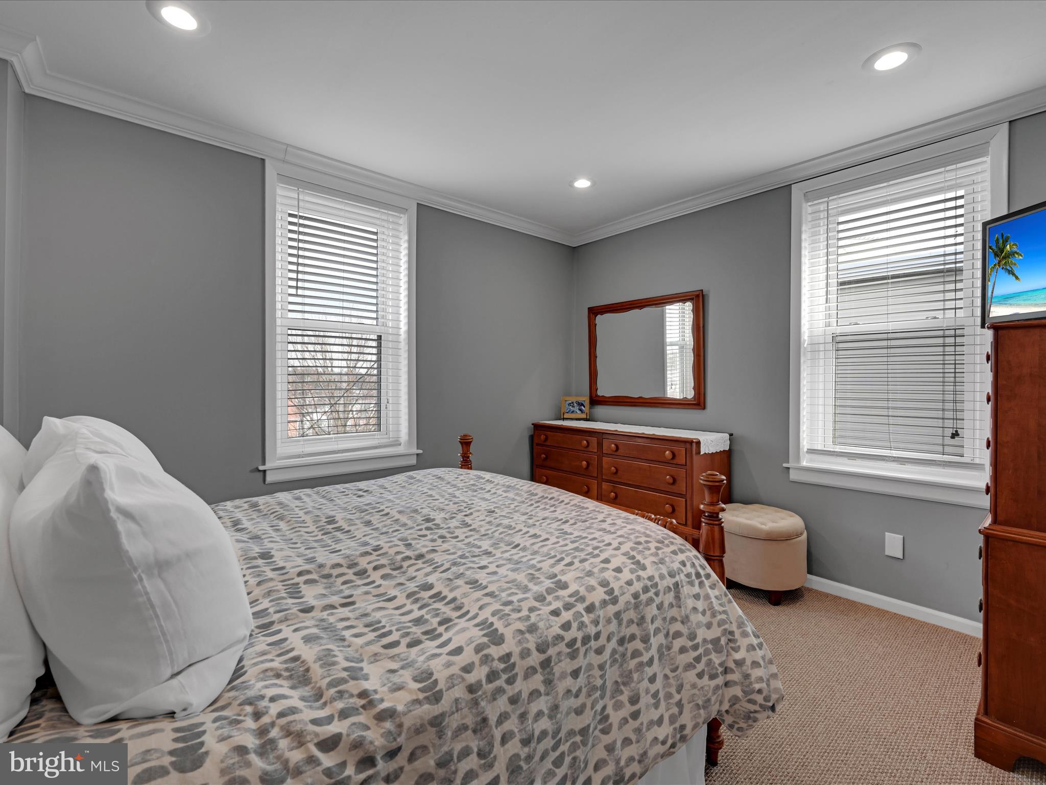 Bedroom with a patterned bedspread, gray walls, and wooden dressers beneath two windows.