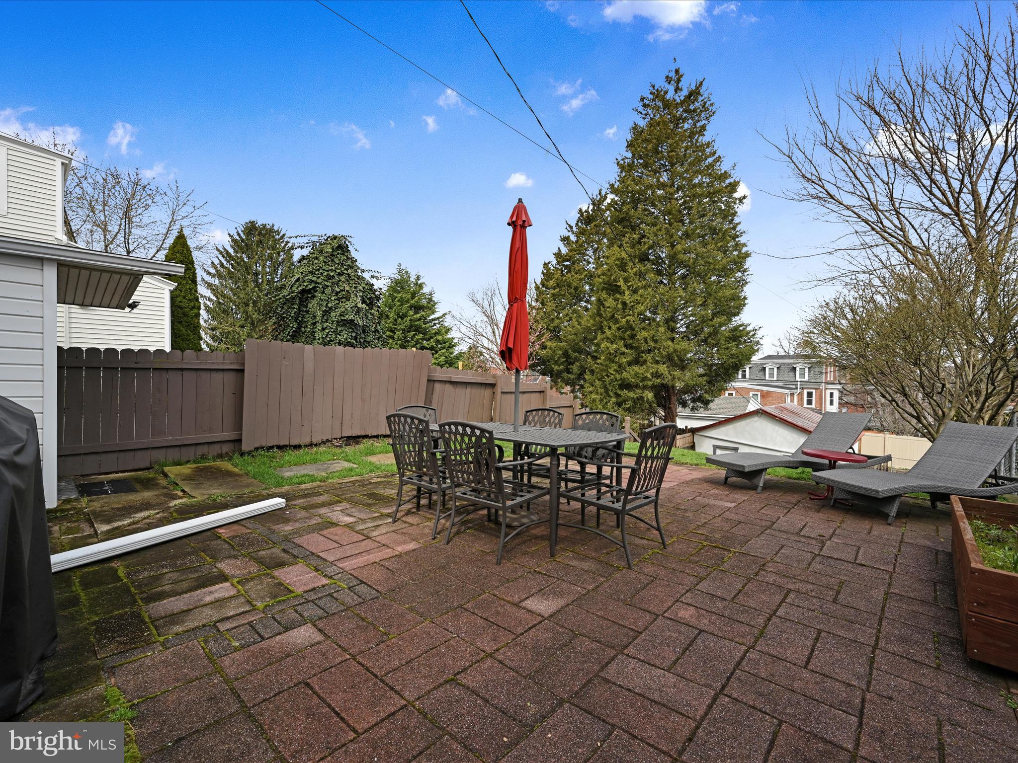 Backyard patio with outdoor dining set, red umbrella, chaise lounges, and privacy fence.