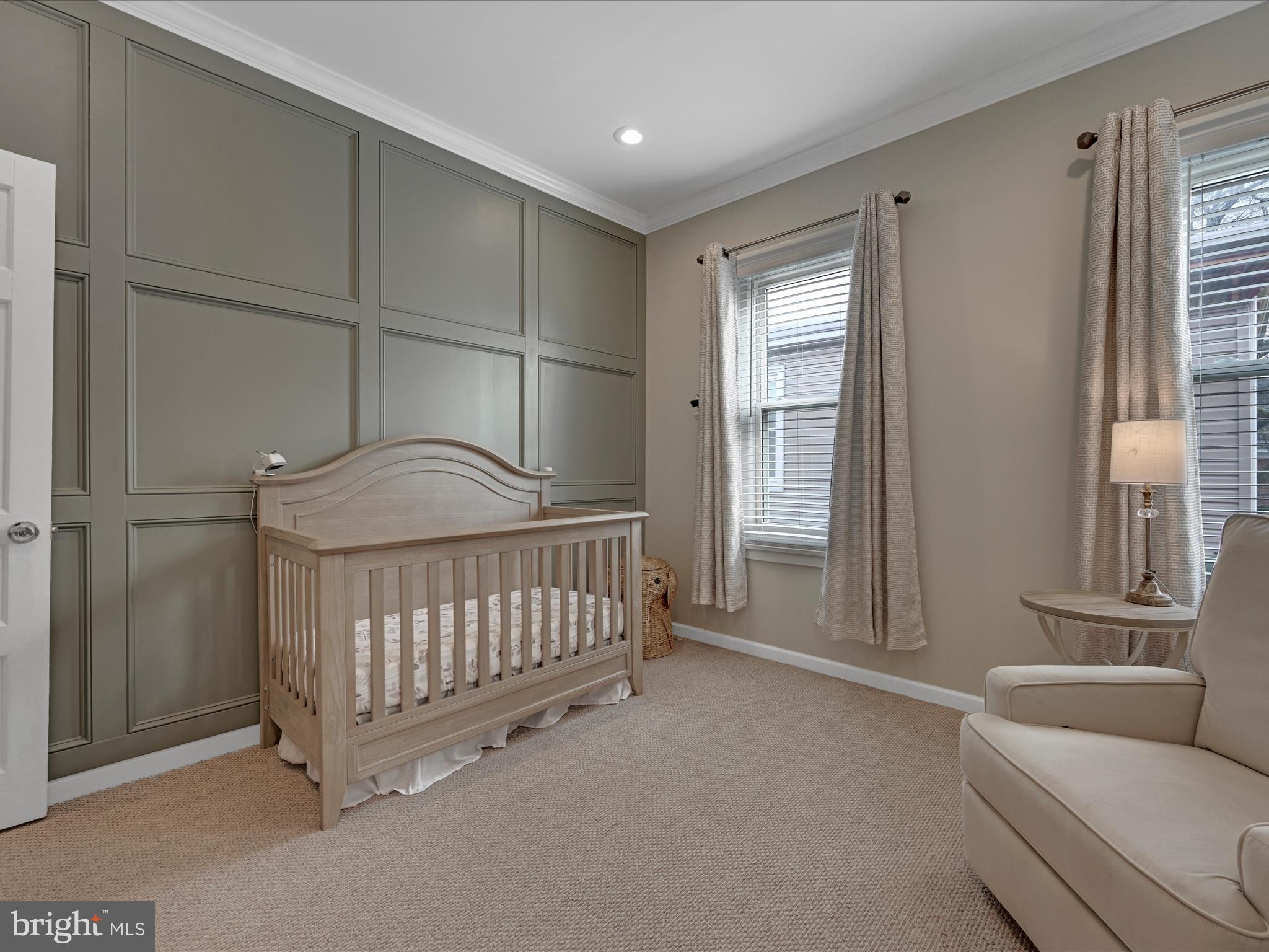 Nursery with a light wood crib, paneled accent wall, curtained windows, and cream armchair.