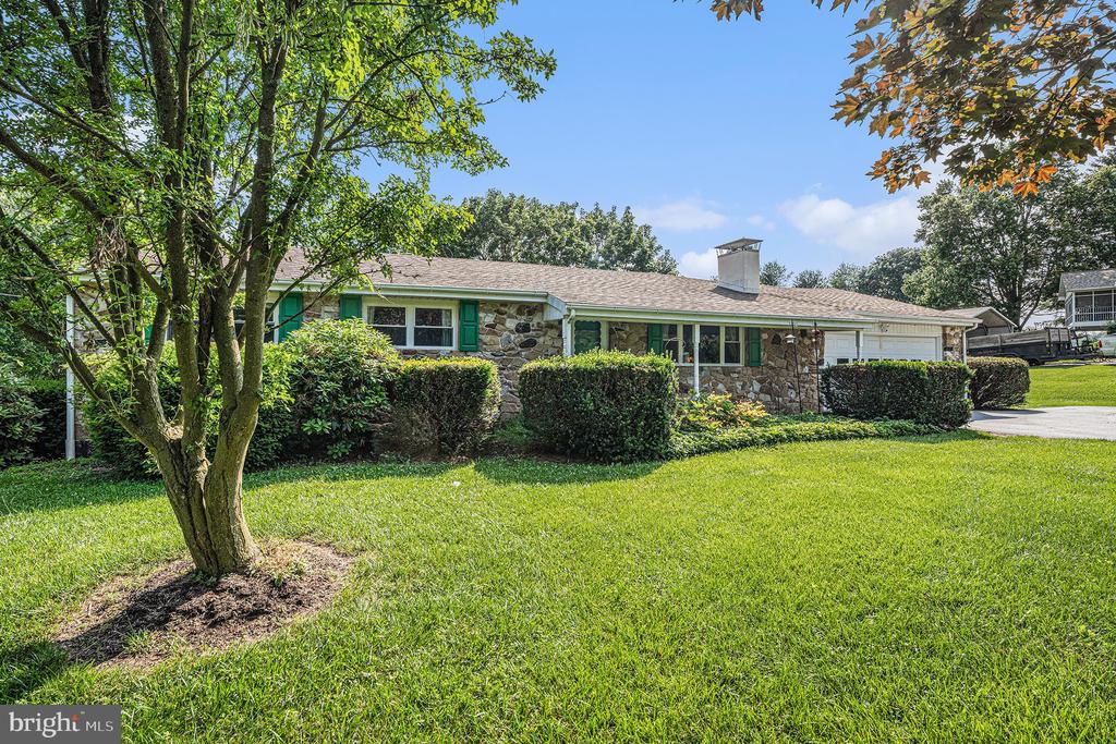 Single-story stone-front home with green shutters, an attached garage, and a tree-framed lawn.