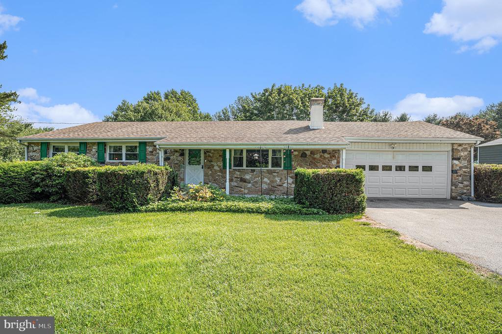 Stone ranch home with green shutters, white garage door, trimmed hedges, and a broad lawn.