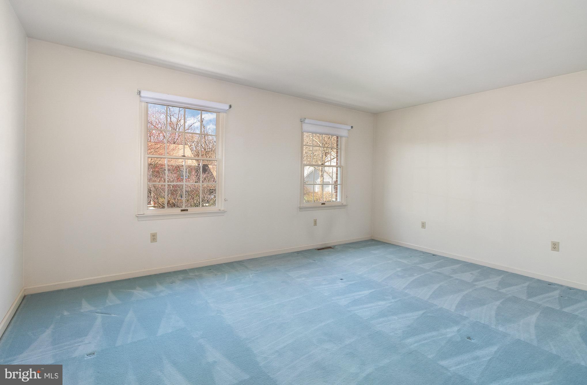 Empty room with blue carpet and two windows overlooking autumn trees.