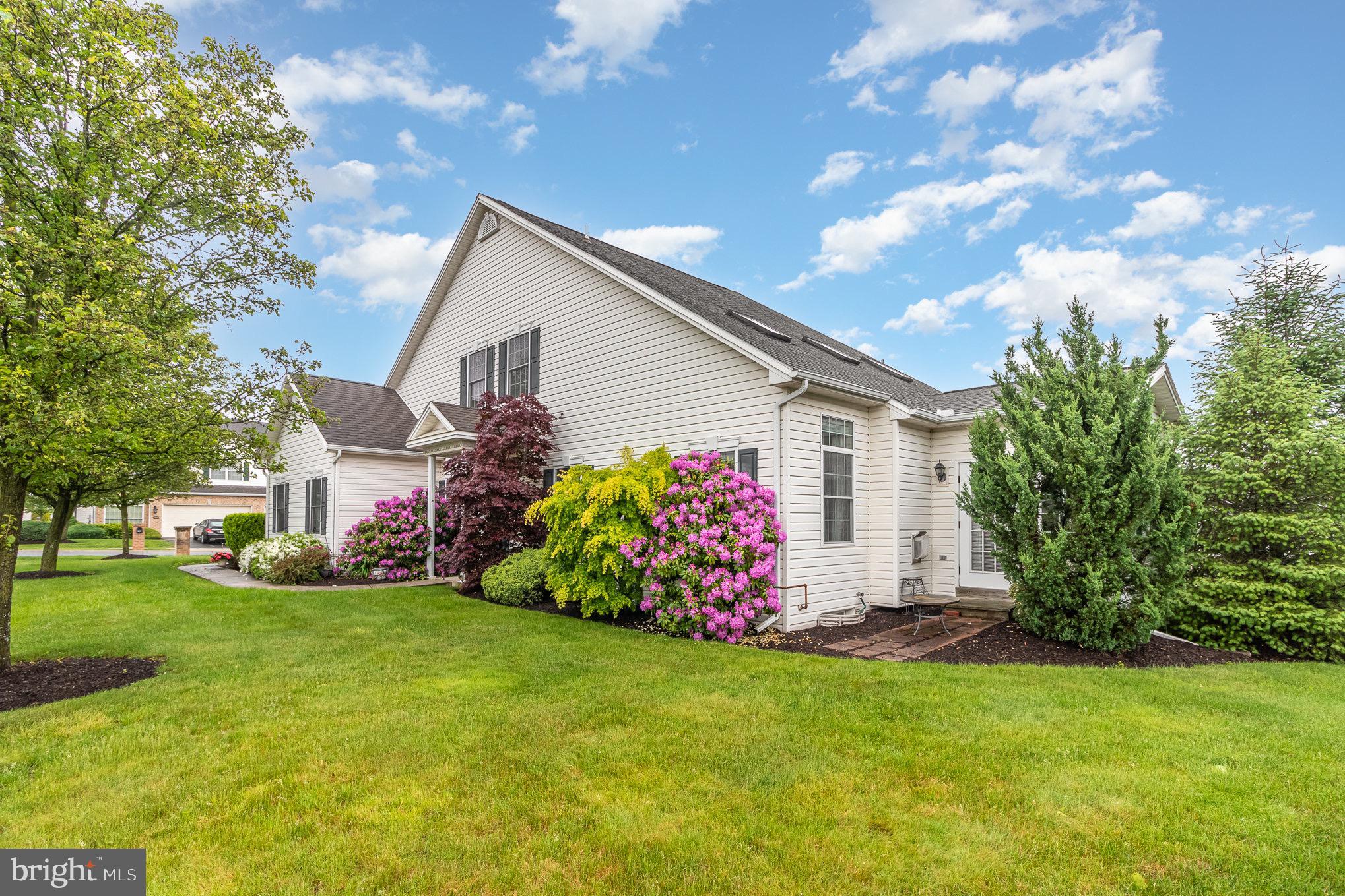 Two-story home with a gabled roof, flowering shrubs, and a small patio beside a broad lawn.