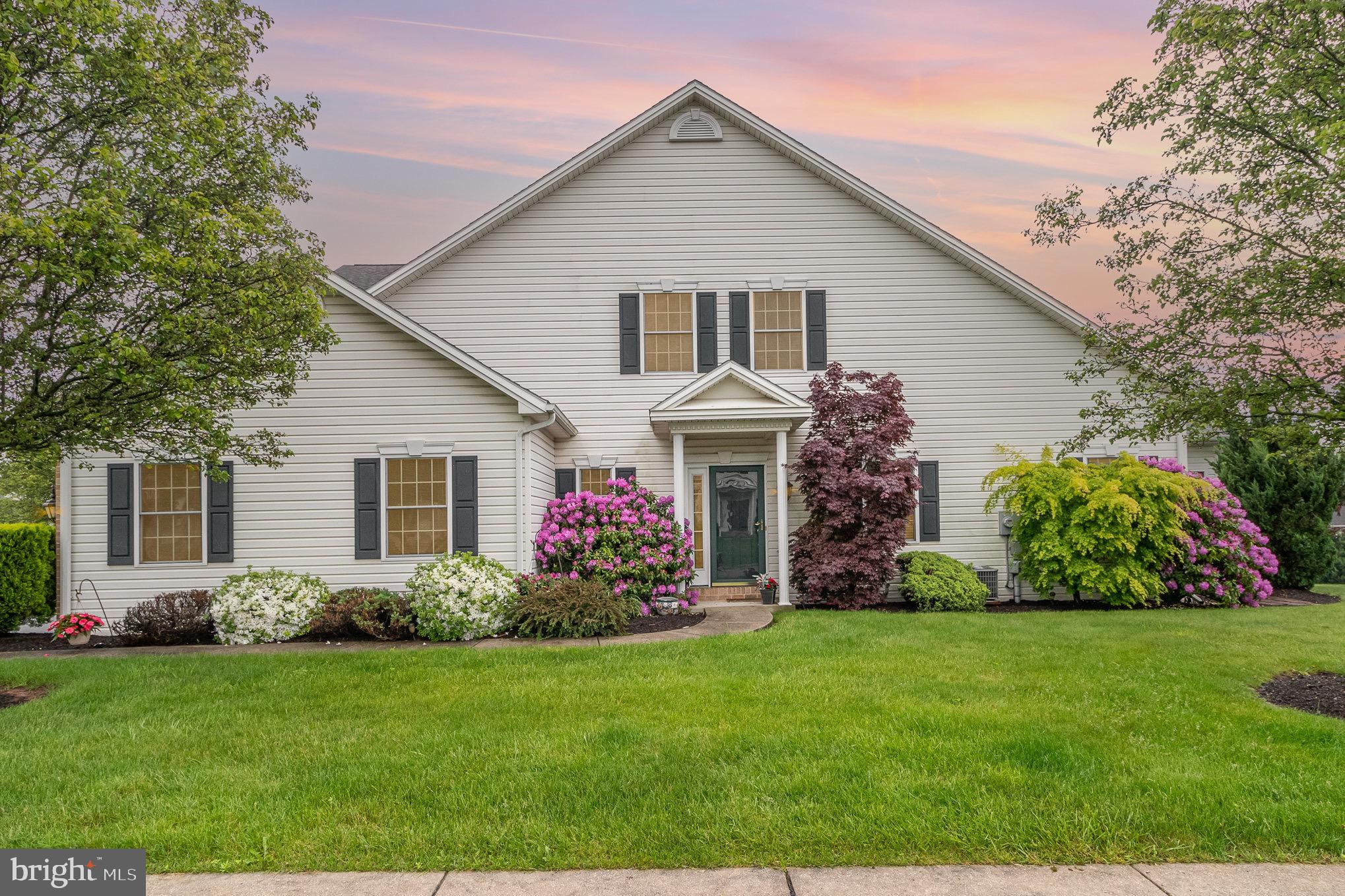 White two-story home with a tall gabled roof, dark shutters, and colorful flowering shrubs along the lawn.