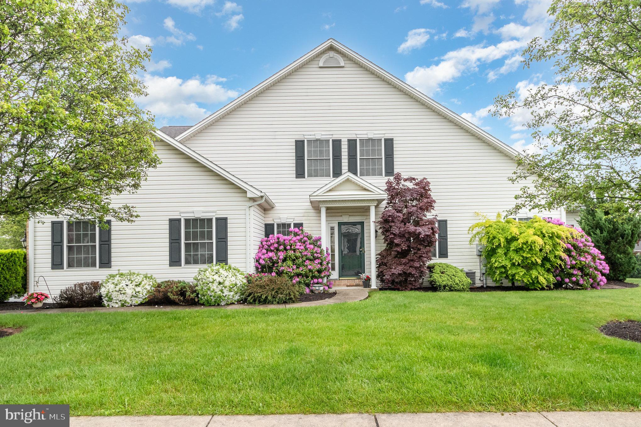 White two-story house with dark shutters, a covered entry, and colorful flowering shrubs across the front lawn.