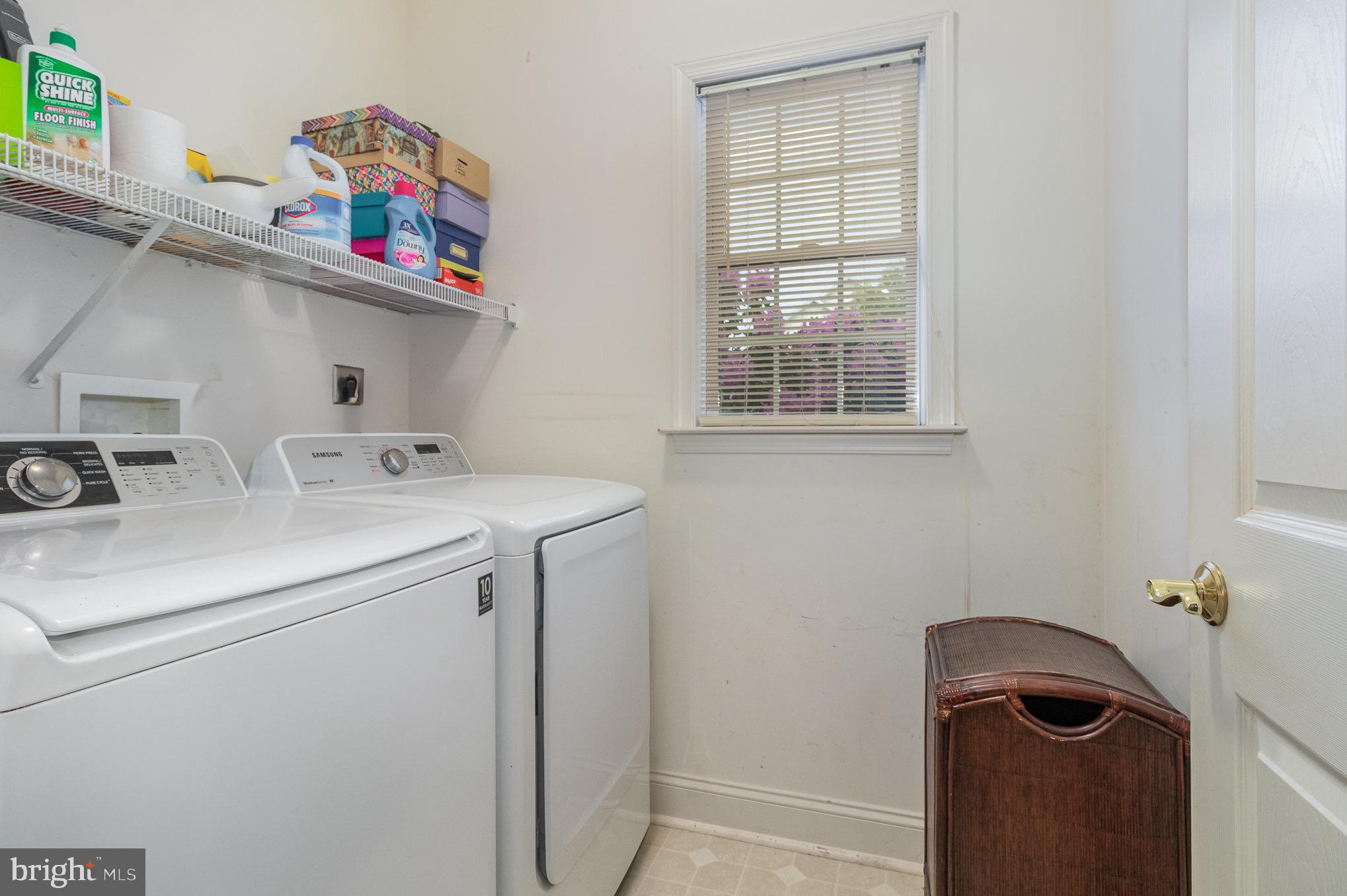 Laundry room with white washer and dryer, storage shelf, window with blinds, and wicker hamper.