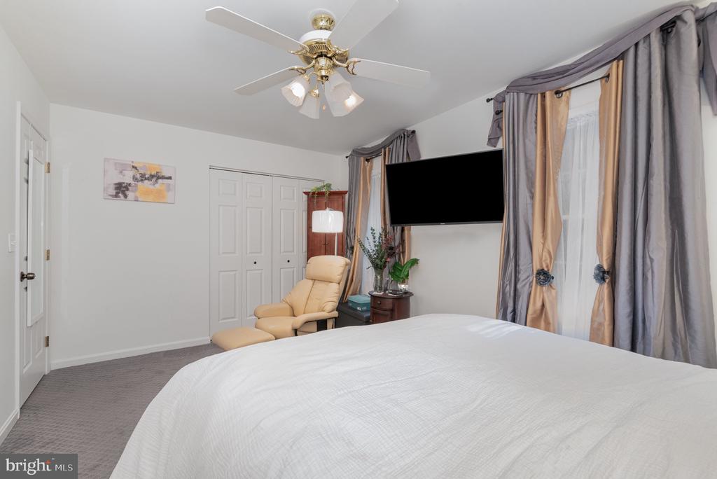 Furnished bedroom with a large bed, beige recliner, ceiling fan, and tall curtained windows.