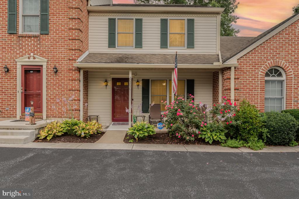 Townhouse front with a red entry door, covered porch, American flag, and flowering shrubs.