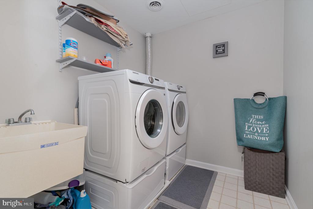 Laundry room with stacked white washer and dryer, utility sink, and shelves above.