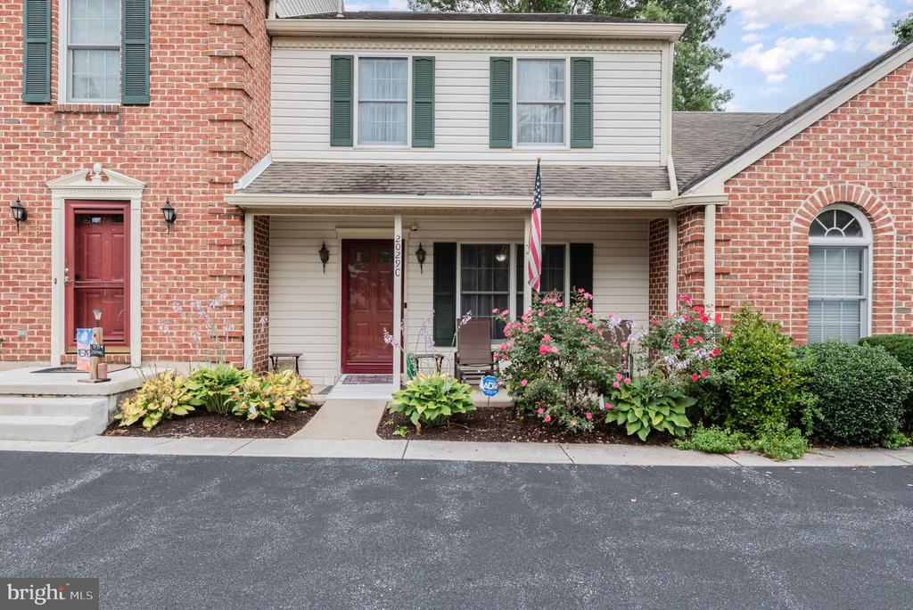 Townhouse exterior with a covered entry, red front door, porch seating, and flowering shrubs.