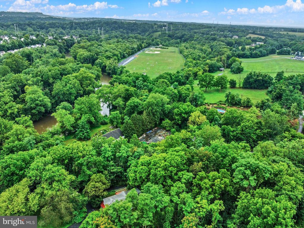 Aerial property view surrounded by trees, with a stream, pool, and open fields.