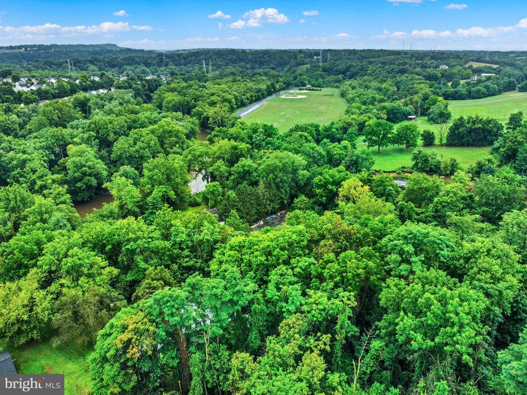 Aerial view of wooded property with a creek, open fields, and sports grounds beyond.