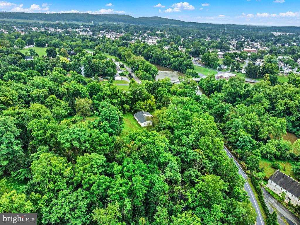 Aerial view of a house amid dense woodland, with a winding road, creek, and distant neighborhood.