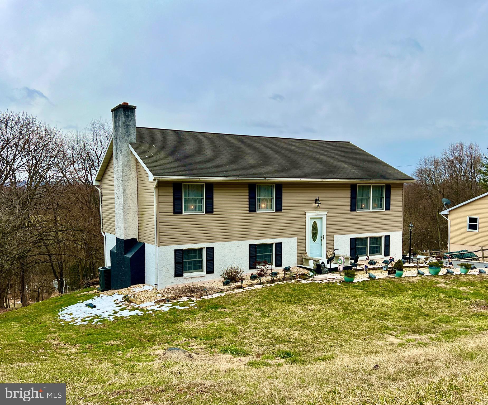 Split-level home with tan siding, a tall stone chimney, and a sloping lawn.